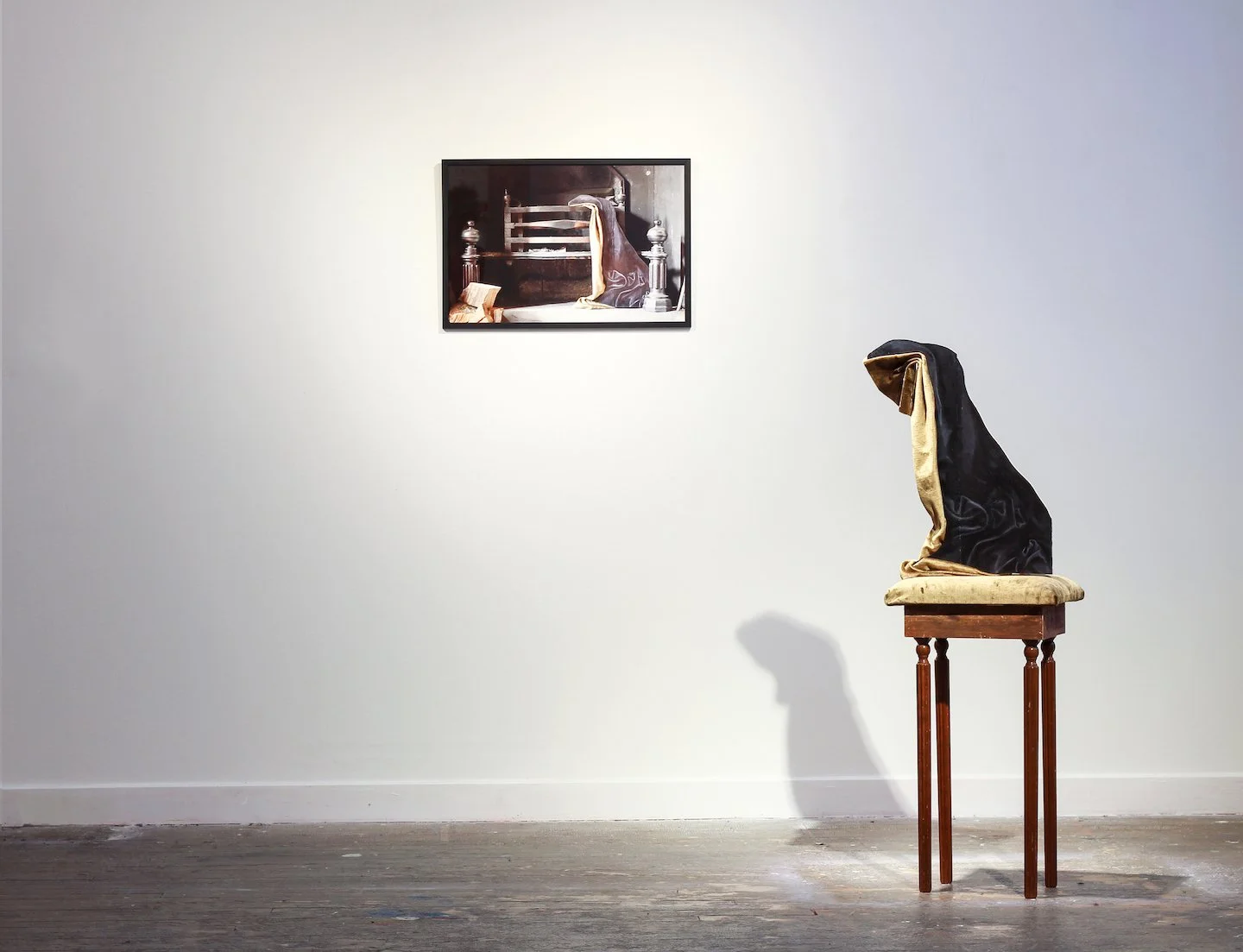 An art installation featuring a wooden table with a velvet black and gold hooded figure sculpture on top, placed in front of a white wall with a framed photograph of a similar hooded figure in the background.
