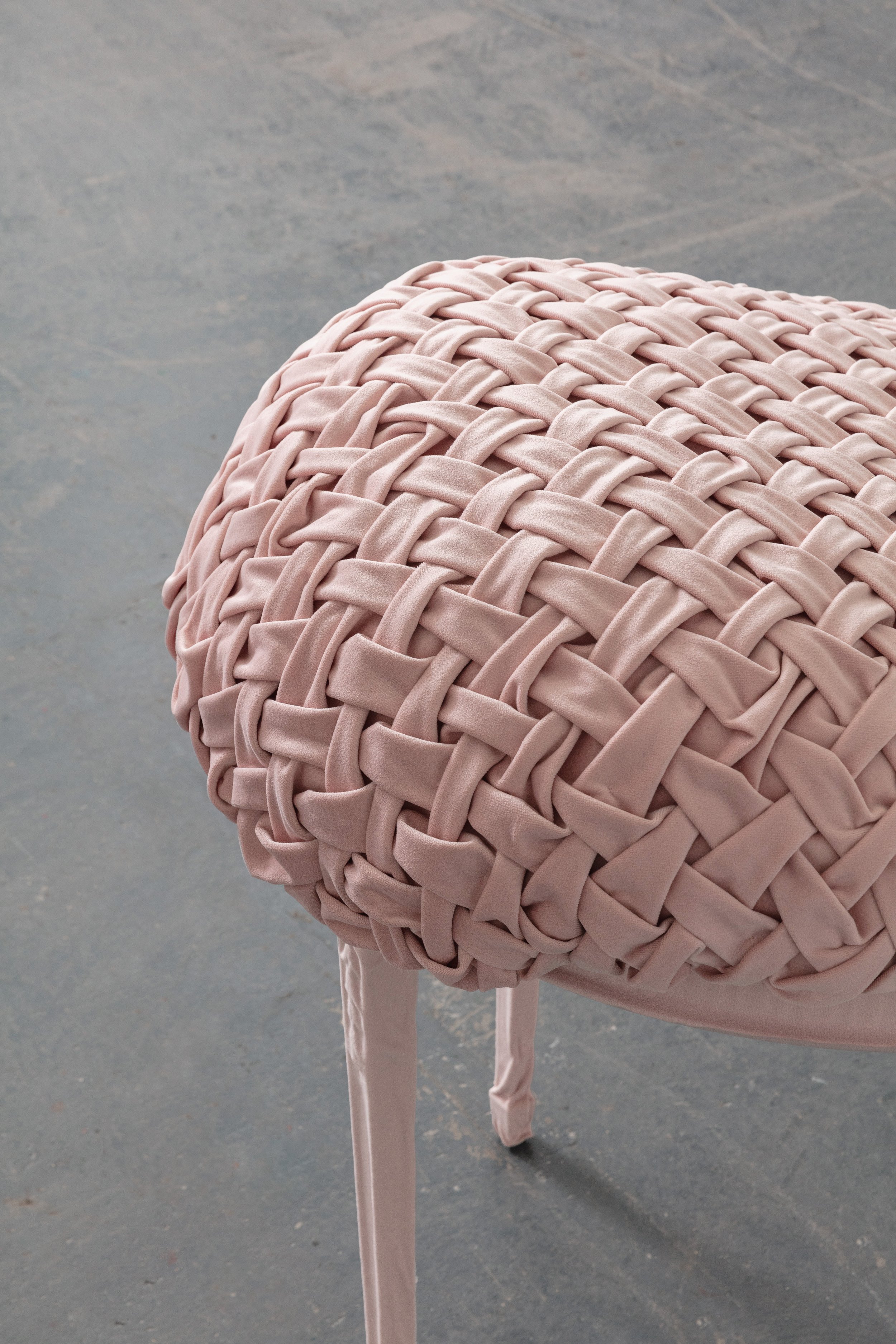A close-up of a round, pink, woven-textured handbag on a stand, with a plain grey background.