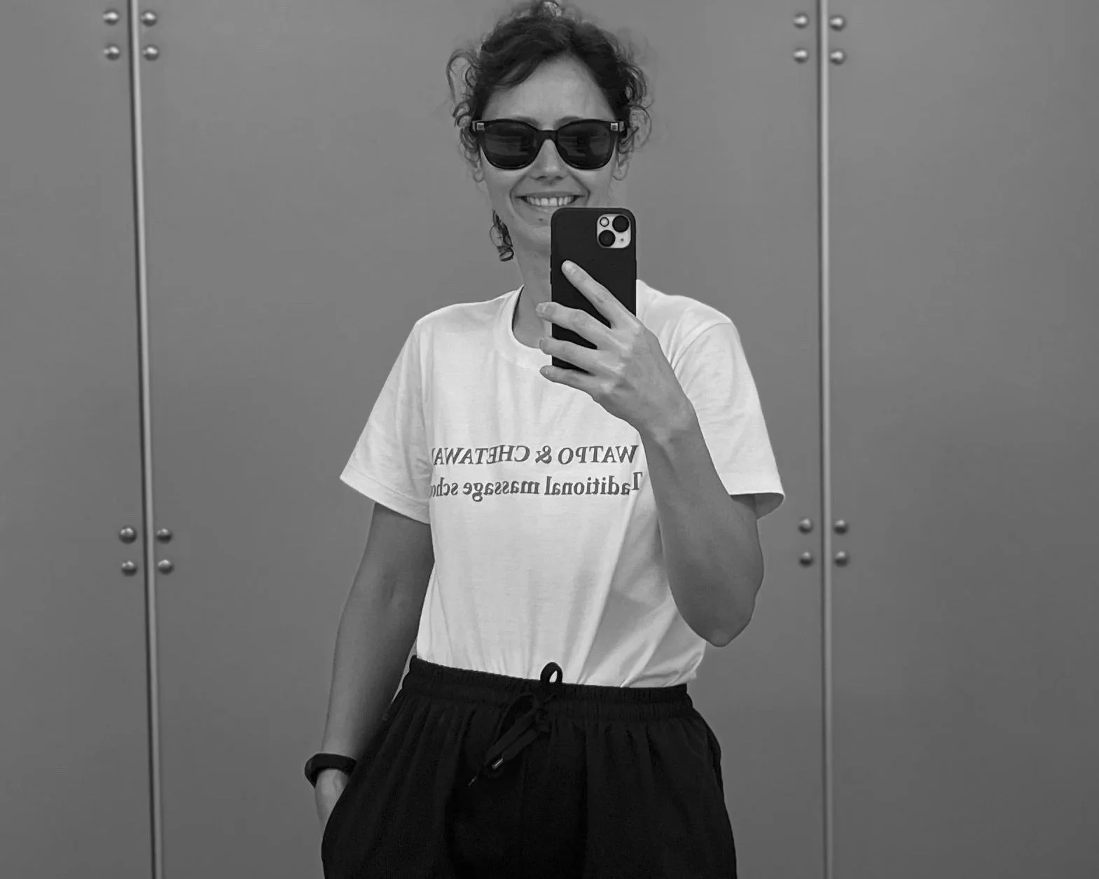 Frau macht Selfie in Aufzug, trägt Sonnenbrille, weißes T-Shirt mit Aufdruck, schwarze Hosen, dunkle Haare, lächelnd.