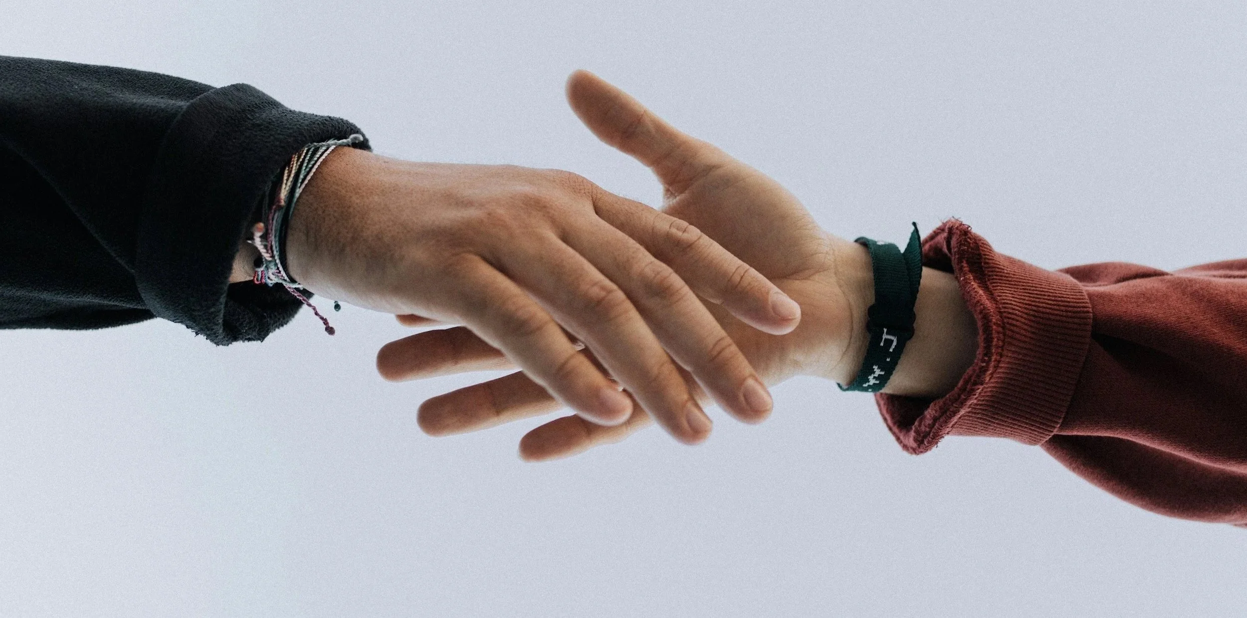 Zwei Menschen greifen sich an den Händen, eine Person trägt eine schwarze Jacke mit Armbändern, die andere eine rote Sweatjacke mit einem grünen Armband.