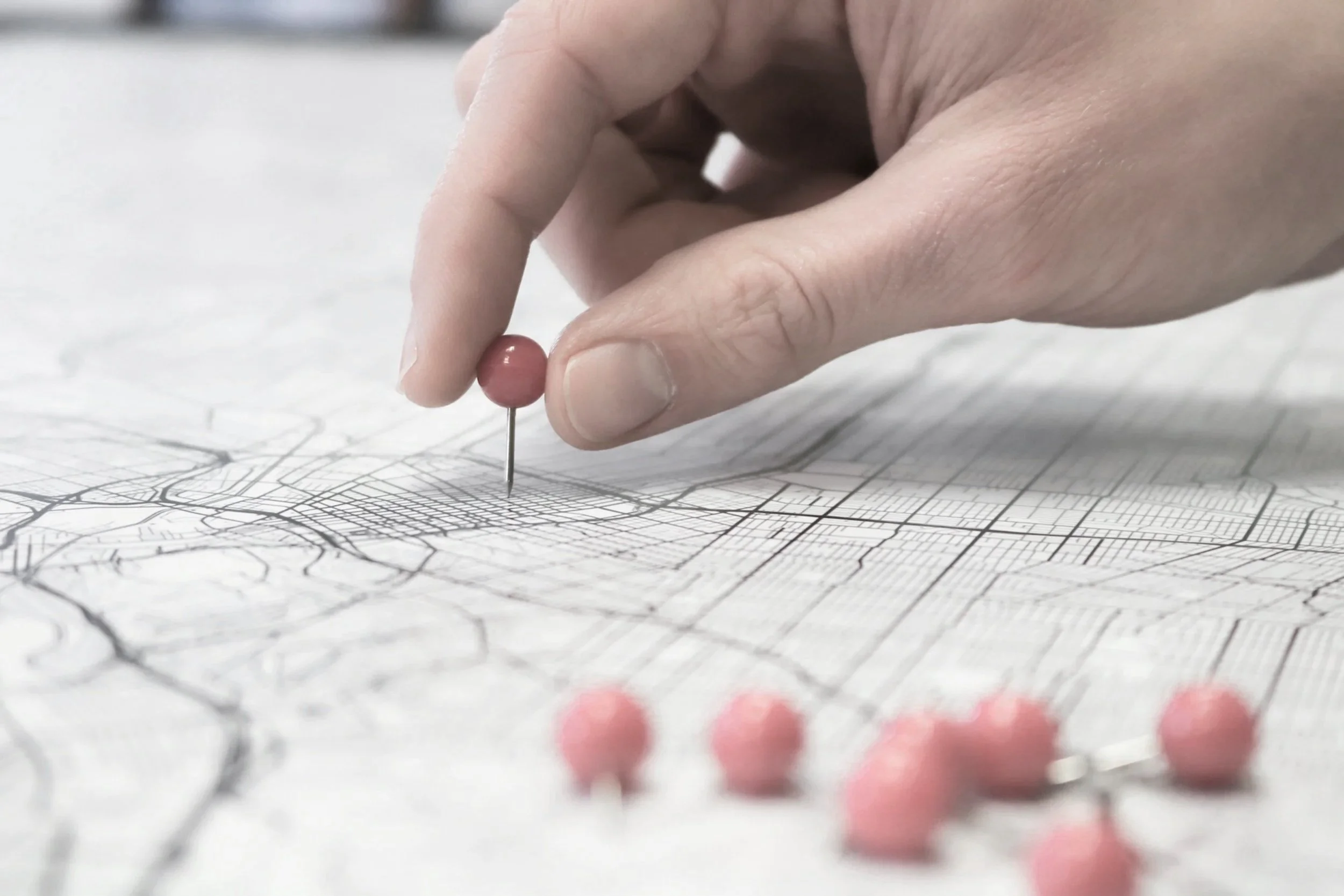 Hand, die eine Nadel mit rosa Kopf in eine Landkarte steckt, um sie zu markieren, mit mehreren rosa Kopf-Nadeln auf der Karte.