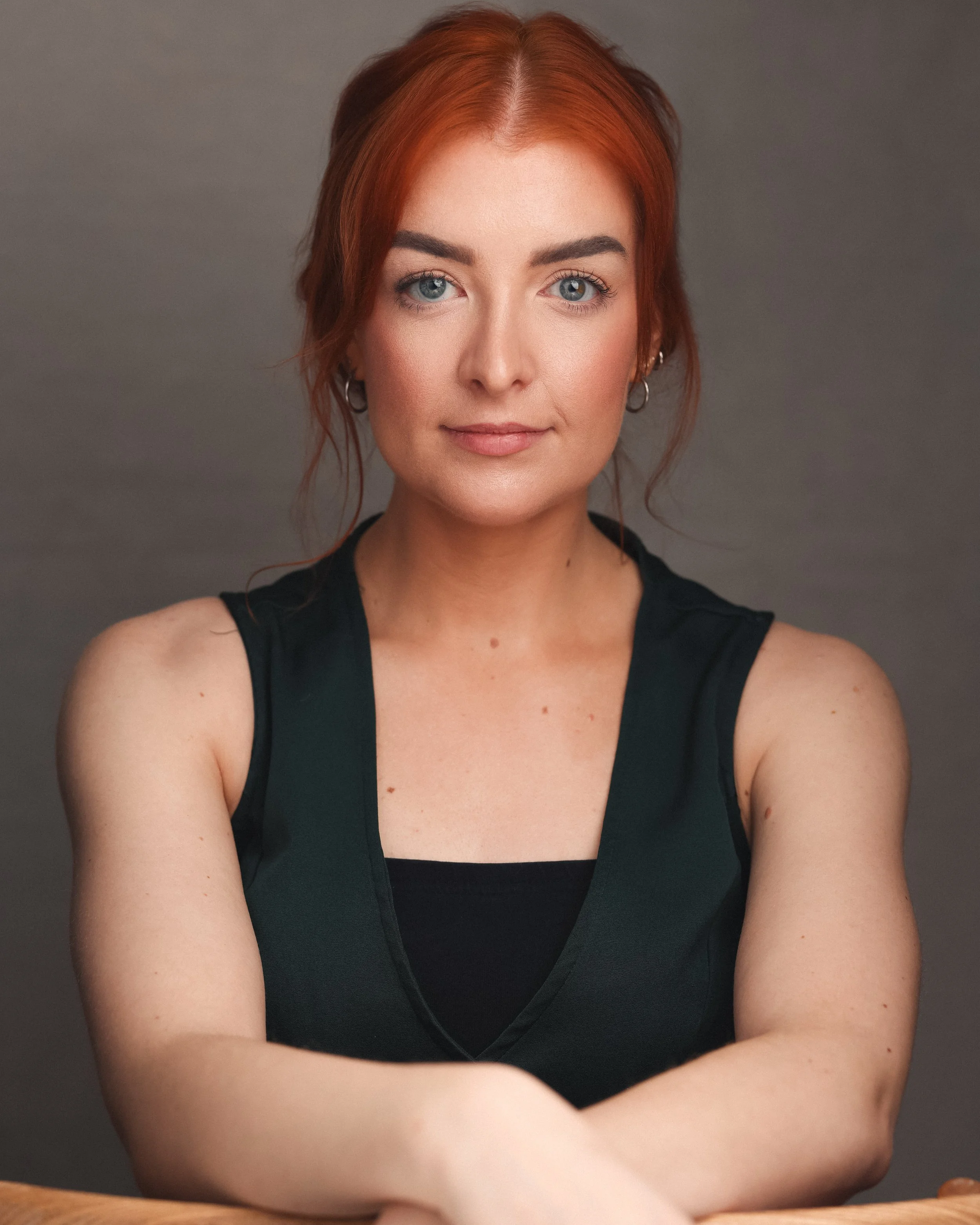 A woman with red hair and blue eyes, wearing a black sleeveless top, sitting with her arms crossed.
