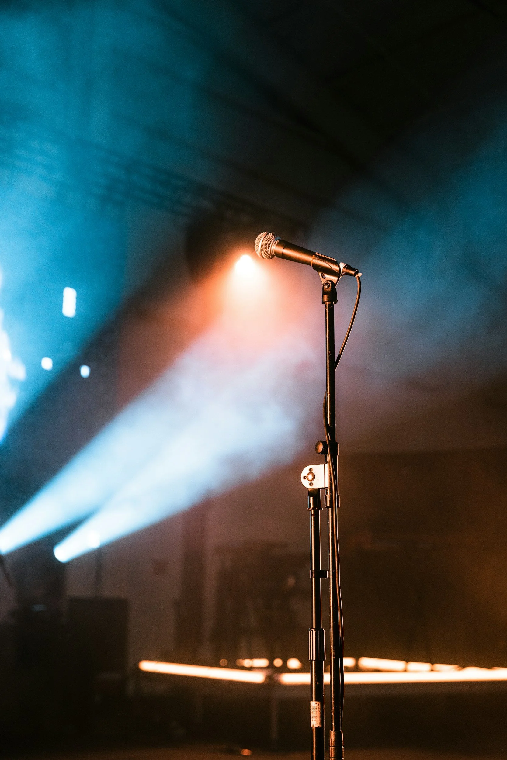 A microphone on a stand in a dimly lit stage or studio, with colorful stage lights in the background creating a moody atmosphere.