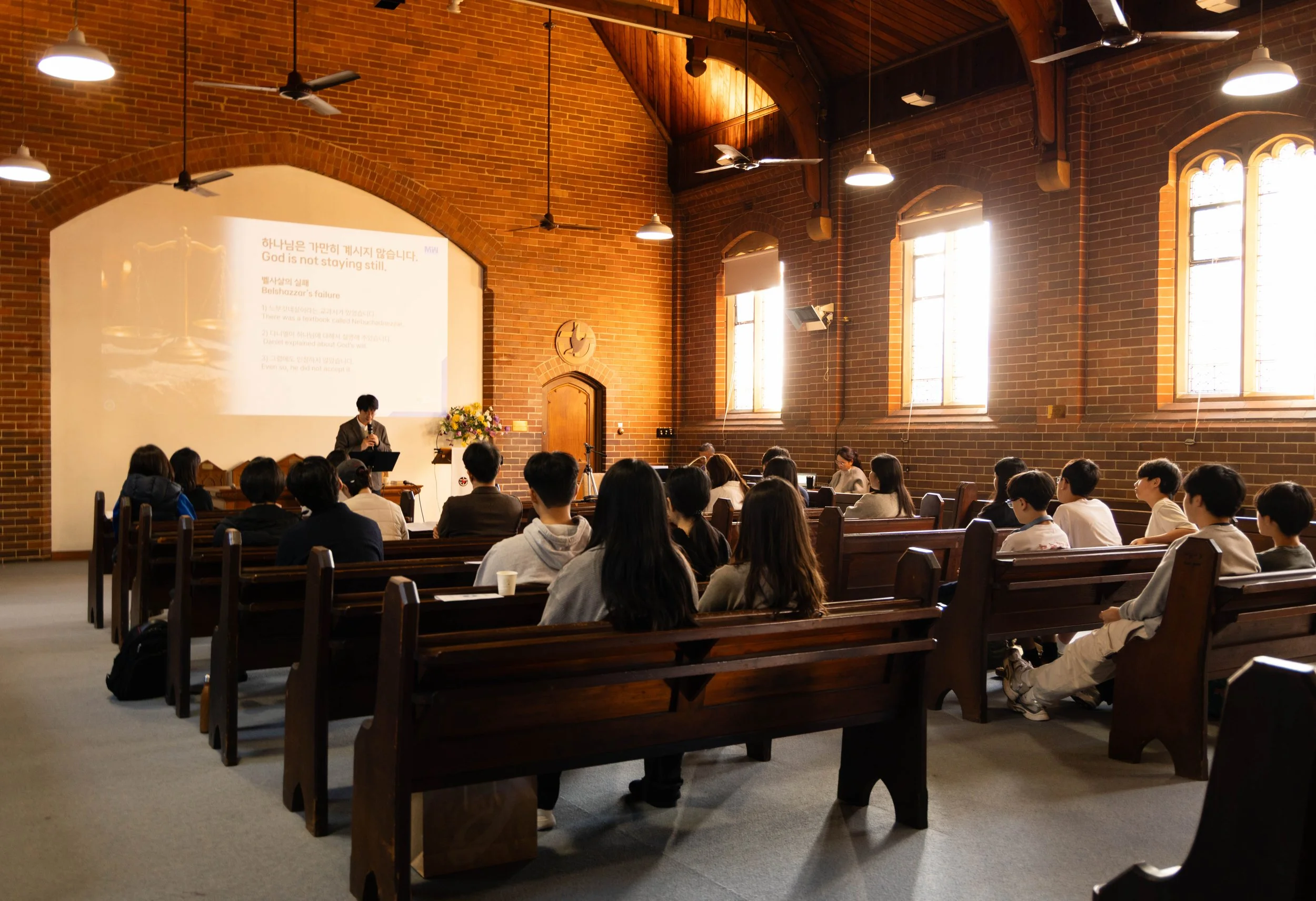 A church or chapel interior with a congregation of people seated on wooden pews, listening to a speaker at a podium. The room has brick walls, large arched windows, and high ceilings with wooden beams and ceiling fans. There is a large screen on one 