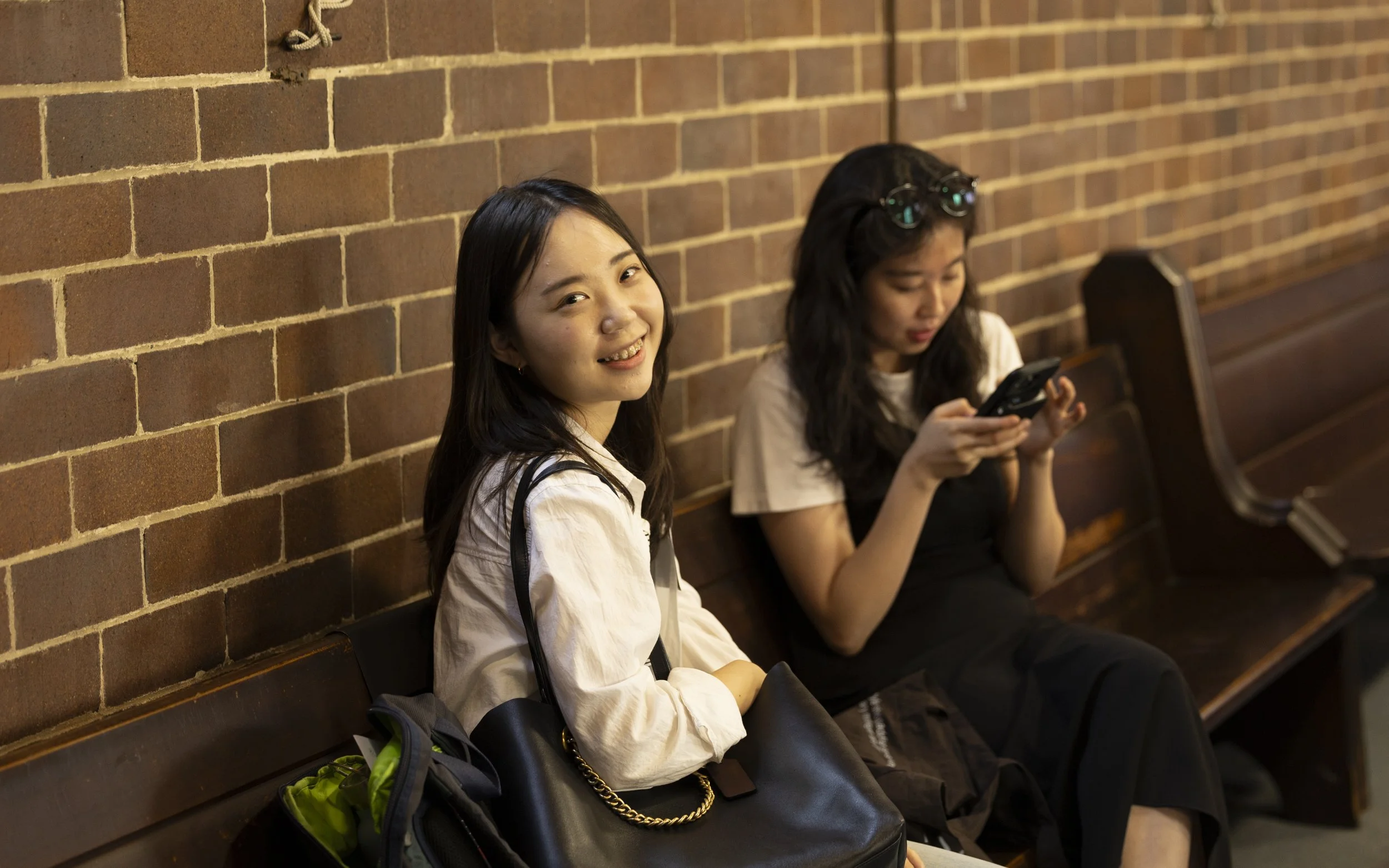 Two young women sitting on a wooden bench against a brick wall. The woman in the foreground has long dark hair, is smiling, and looking at the camera. She is wearing a white shirt and has a black bag with a chain strap. The woman in the background is