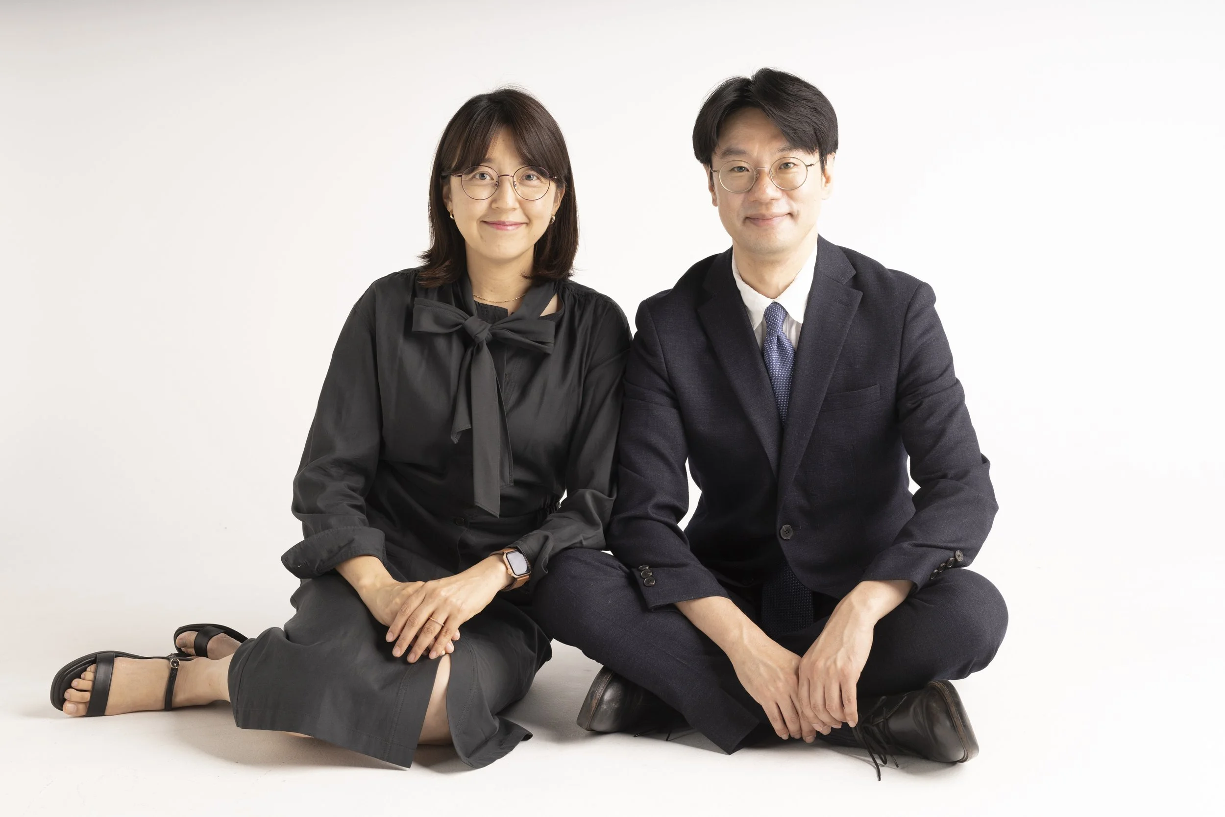 A man and a woman sitting on the floor against a plain white background. The woman is wearing glasses, a black blouse with a bow, and black skirt, while the man is dressed in a dark suit with a white shirt and tie. They are smiling at the camera.