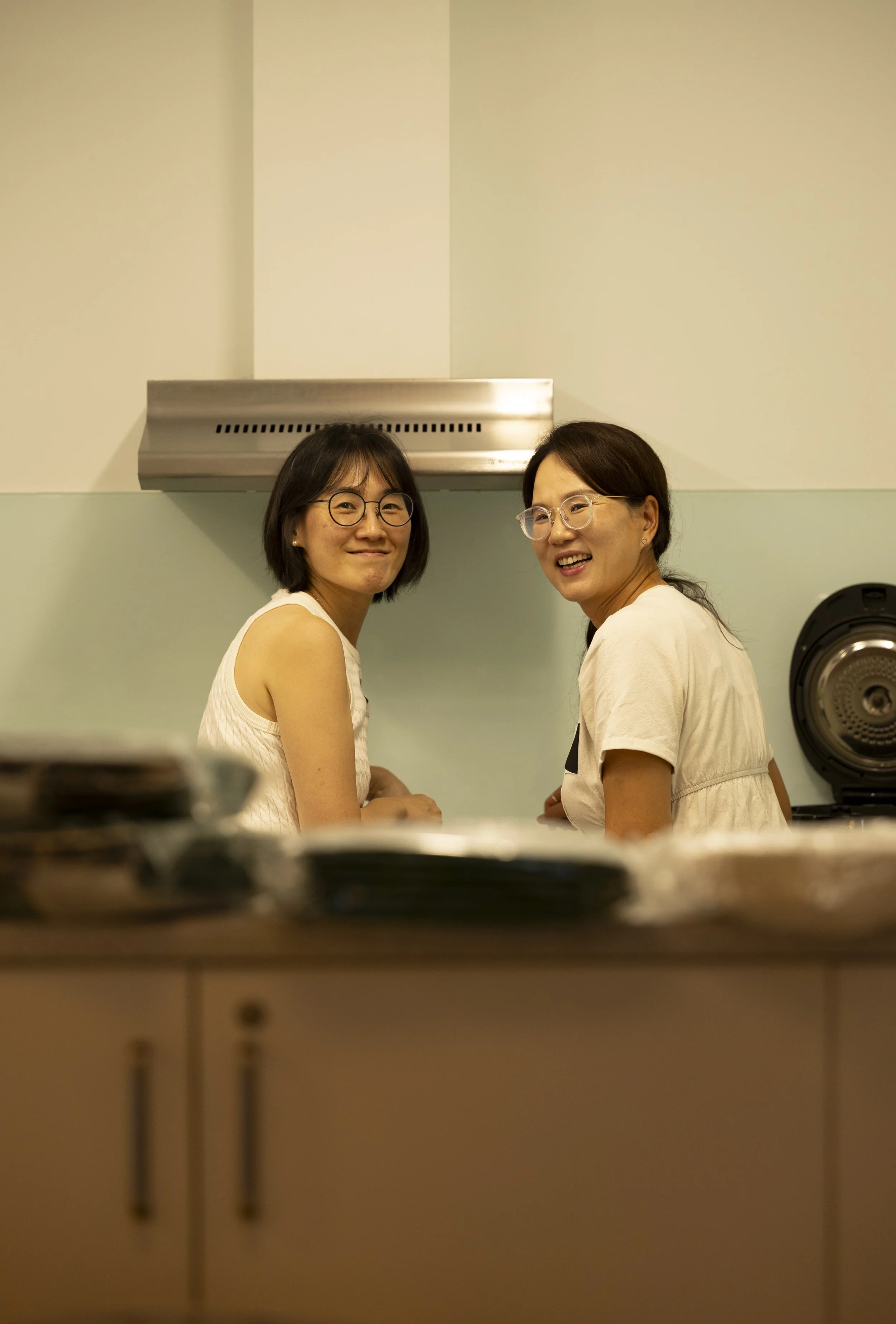 Two women smiling and looking toward the camera in a kitchen, with a stove hood and a speaker in the background, viewed from behind a counter with dishes in the foreground.