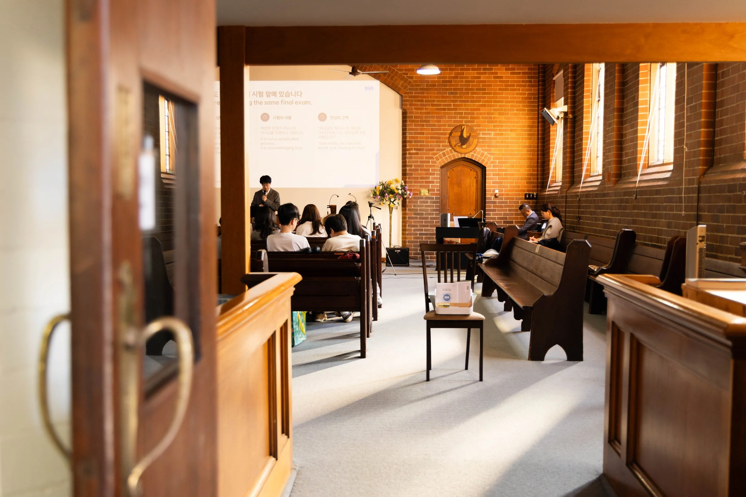 The image shows the interior of a church or chapel with a group of people sitting in pews. There is a woman standing at the front with a microphone, possibly giving a sermon or speech. The church has brick walls, tall windows, and a large projected screen displaying text at the front. The photo is taken from a side aisle, with the door frame partially visible in the foreground.