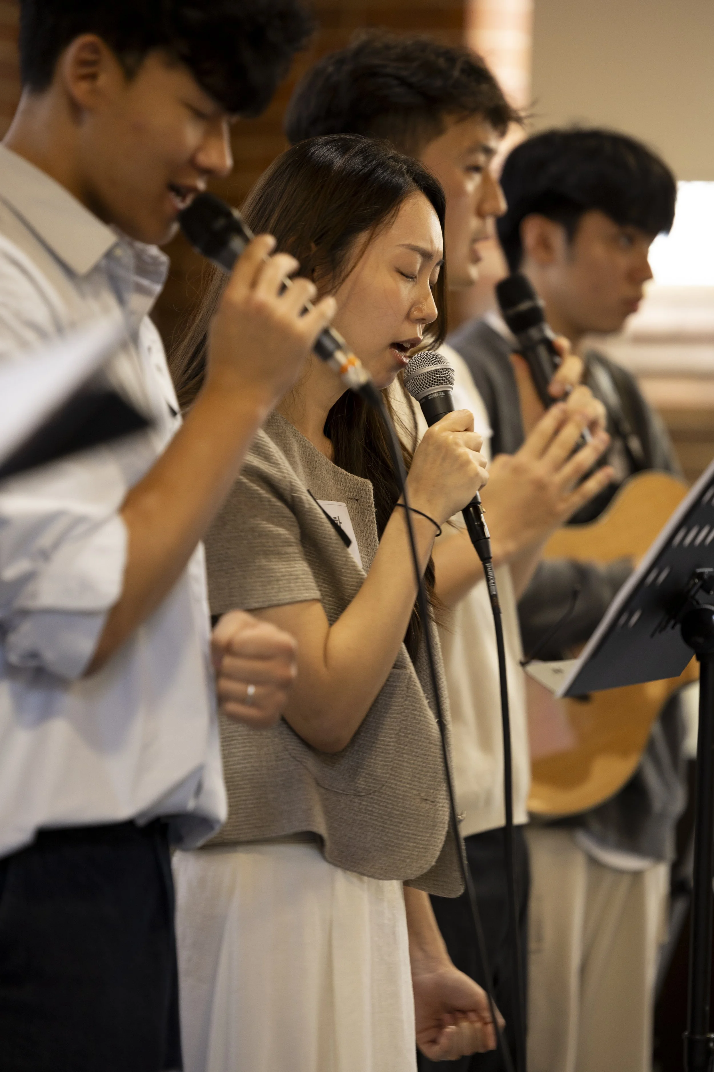 A group of people standing in a line, holding microphones, with head bowed and eyes closed, appearing to be engaged in a prayer or moment of reflection.