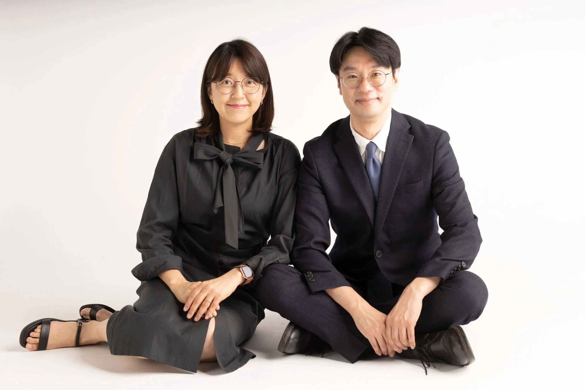 A woman and a man sitting on the floor against a white background. The woman is wearing glasses, a black blouse, and sandals, smiling with her left hand resting on her knee. The man is wearing glasses, a dark suit with a white shirt and tie, smiling with his hands resting on his knees.