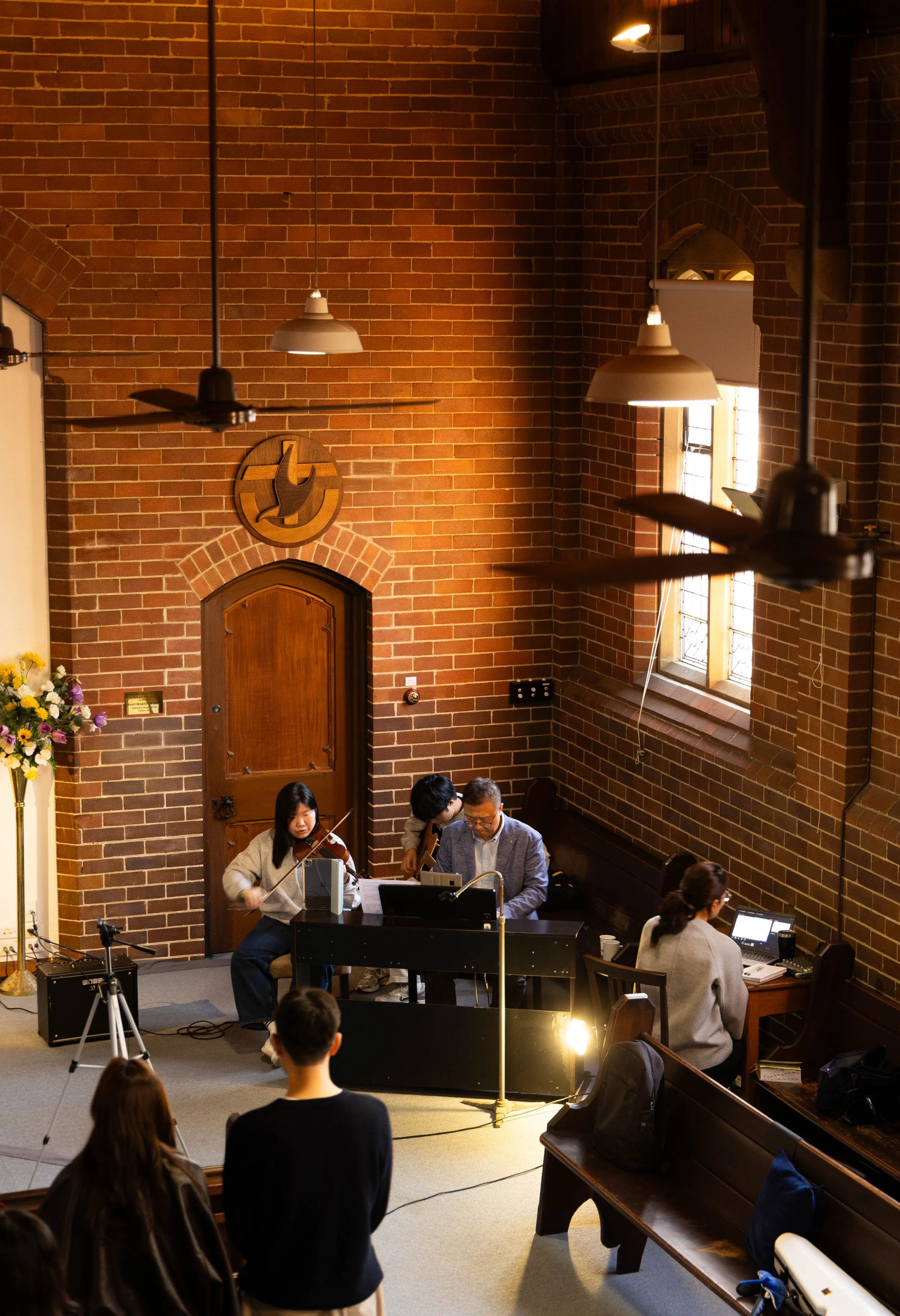 People performing music in a church or concert hall with brick walls, a wooden door, and stained glass windows.