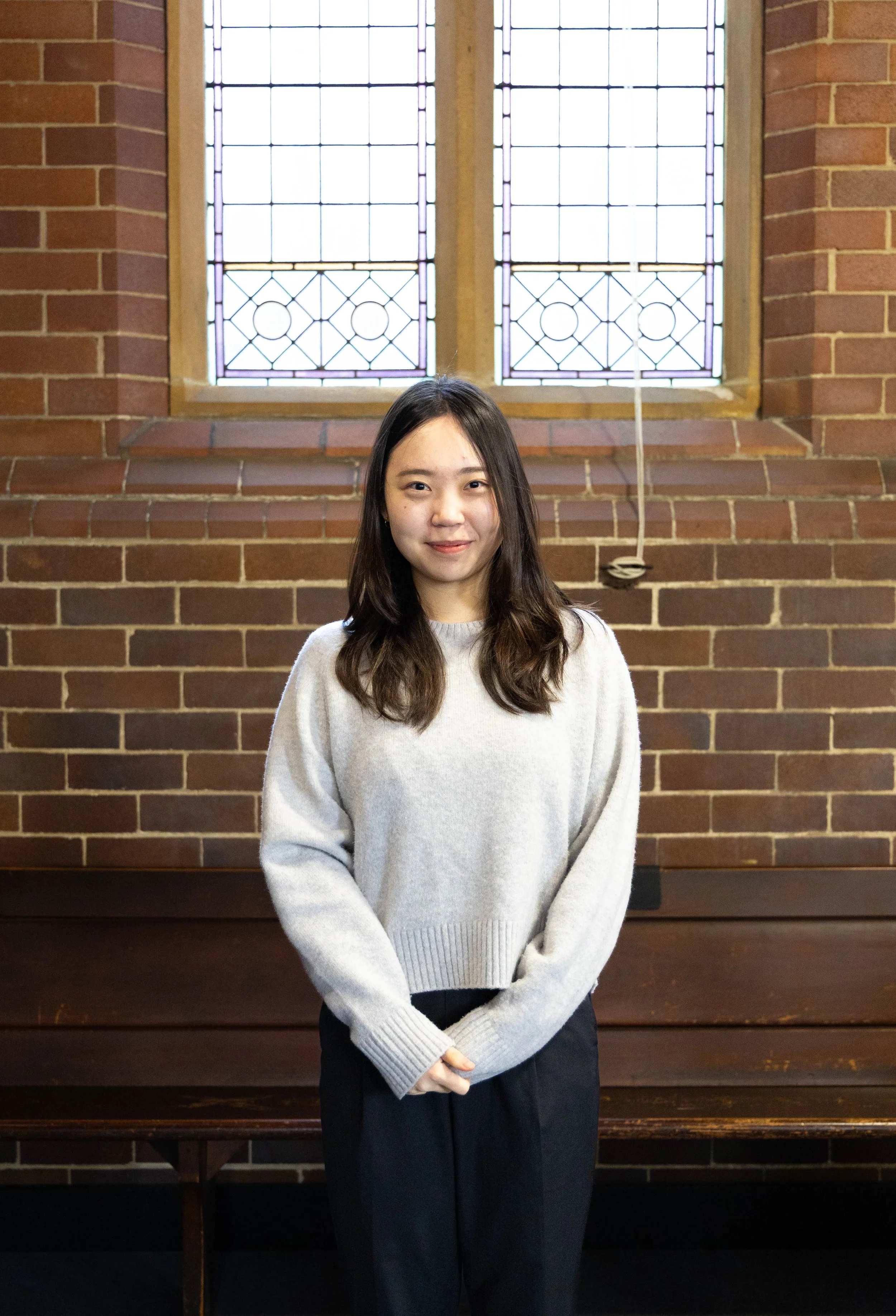 A young woman with long dark hair and light skin, wearing a light gray sweater and dark pants, standing indoors in front of a brick wall with two stained glass windows.