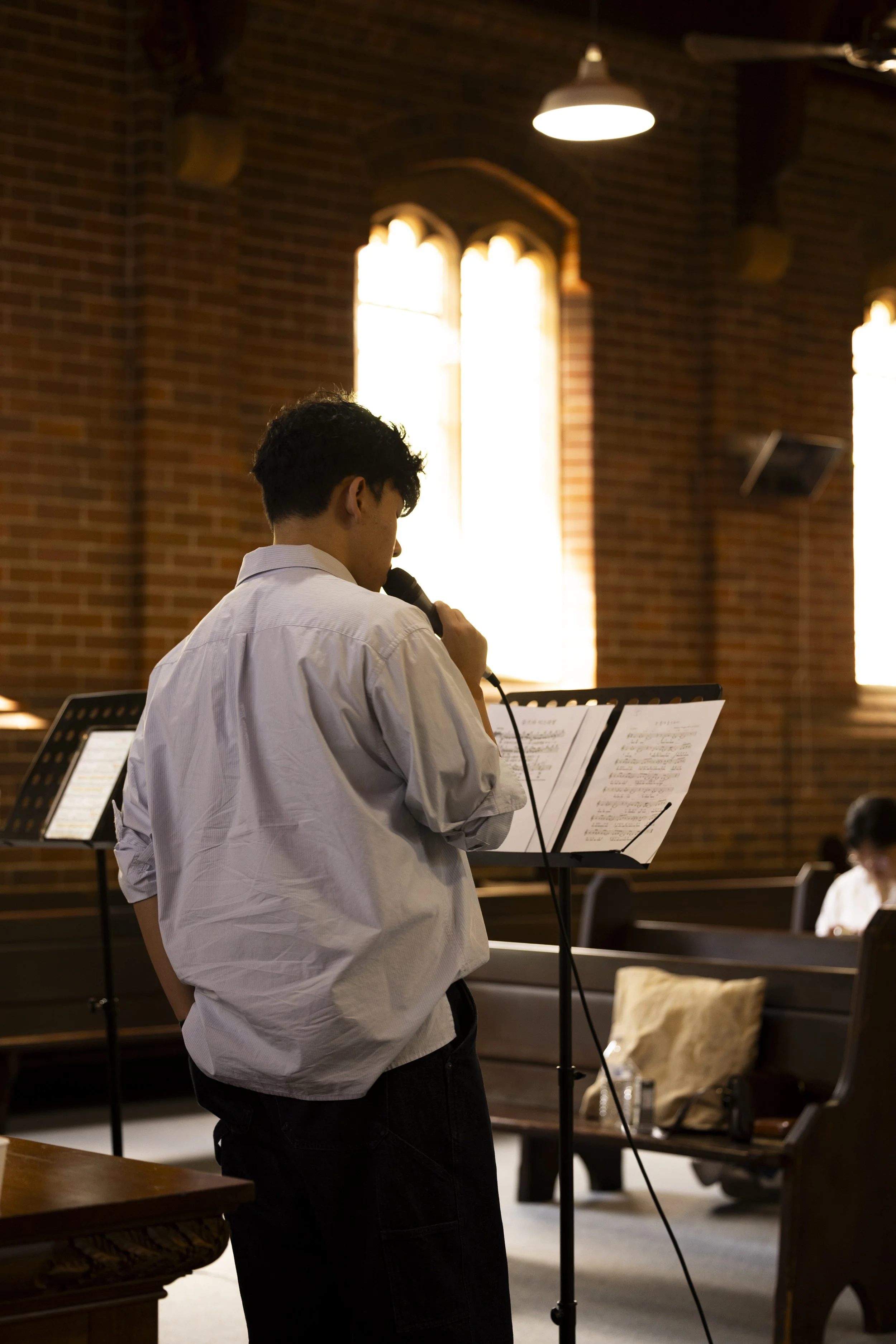A young man in a white shirt and black pants standing and singing into a microphone in a church or concert hall with stained glass windows and music stands.