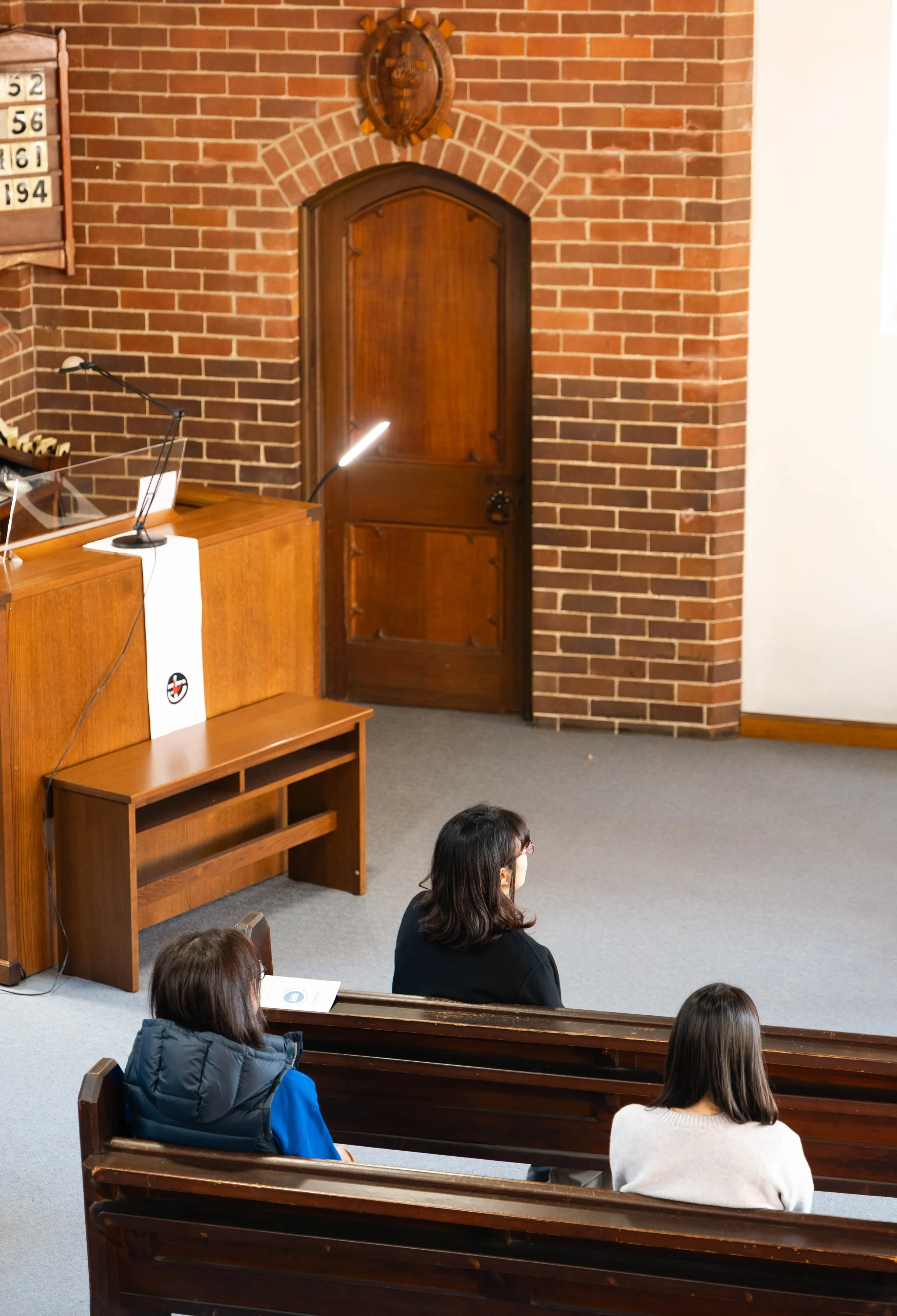 People seated in church pews attending a service, with brick walls and a wooden door in the background.