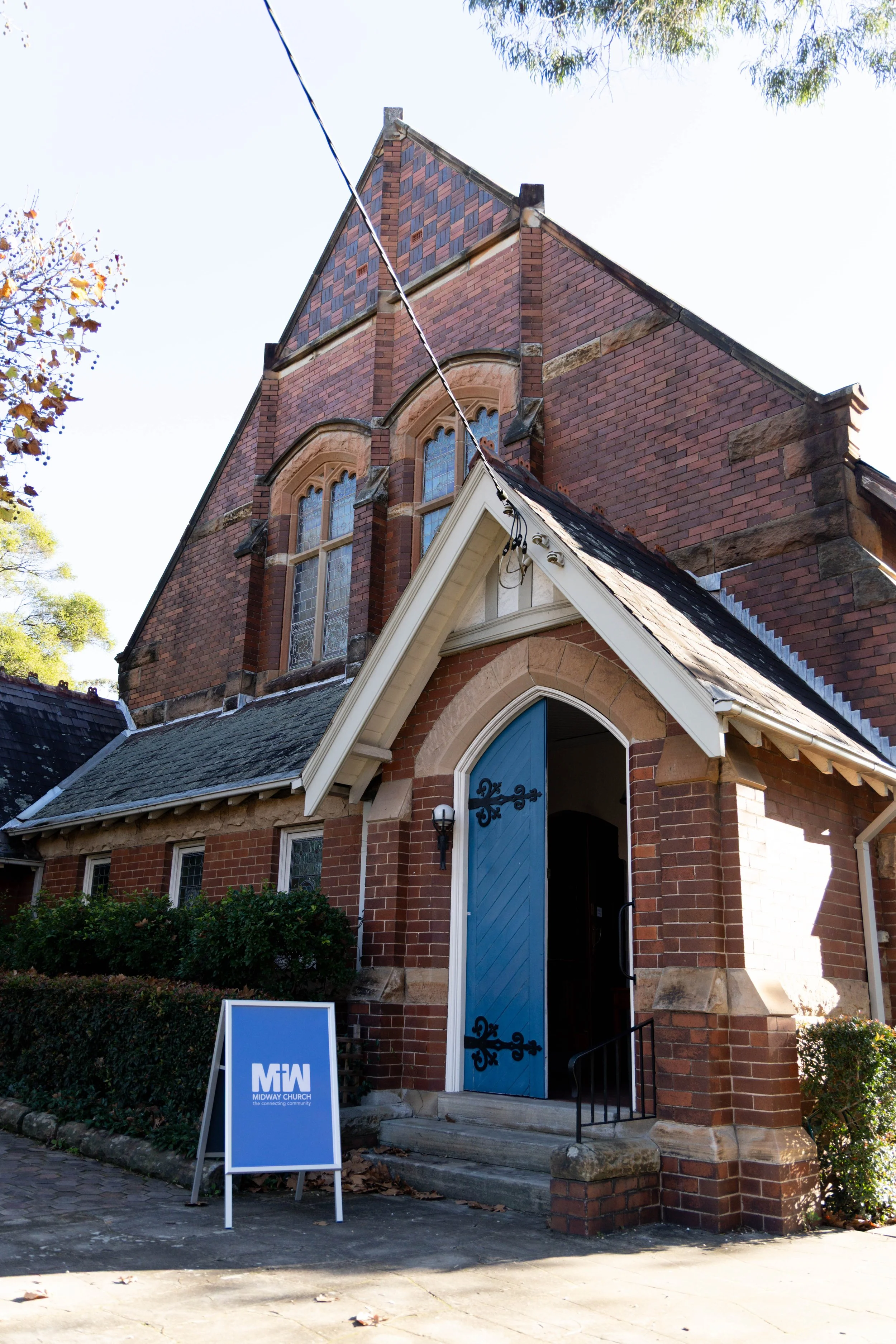 Exterior view of Midway Church, a red brick church building with arched stained glass windows, a light blue door, and a sign in front that reads "MIDWAY CHURCH connecting community." There are trees and a clear sky in the background.