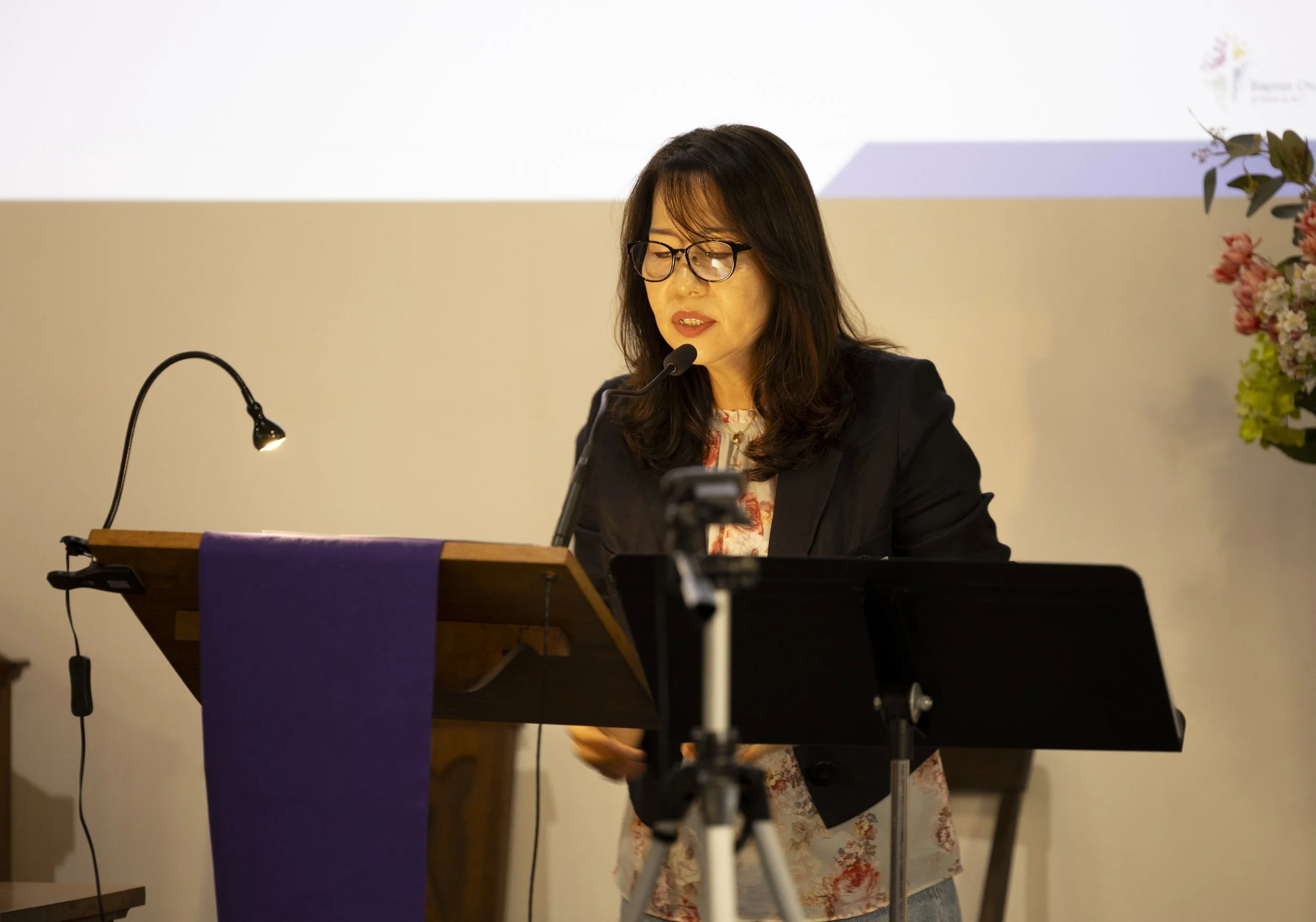 Woman with glasses reading from a paper at a podium, with flowers on the side and a microphone.