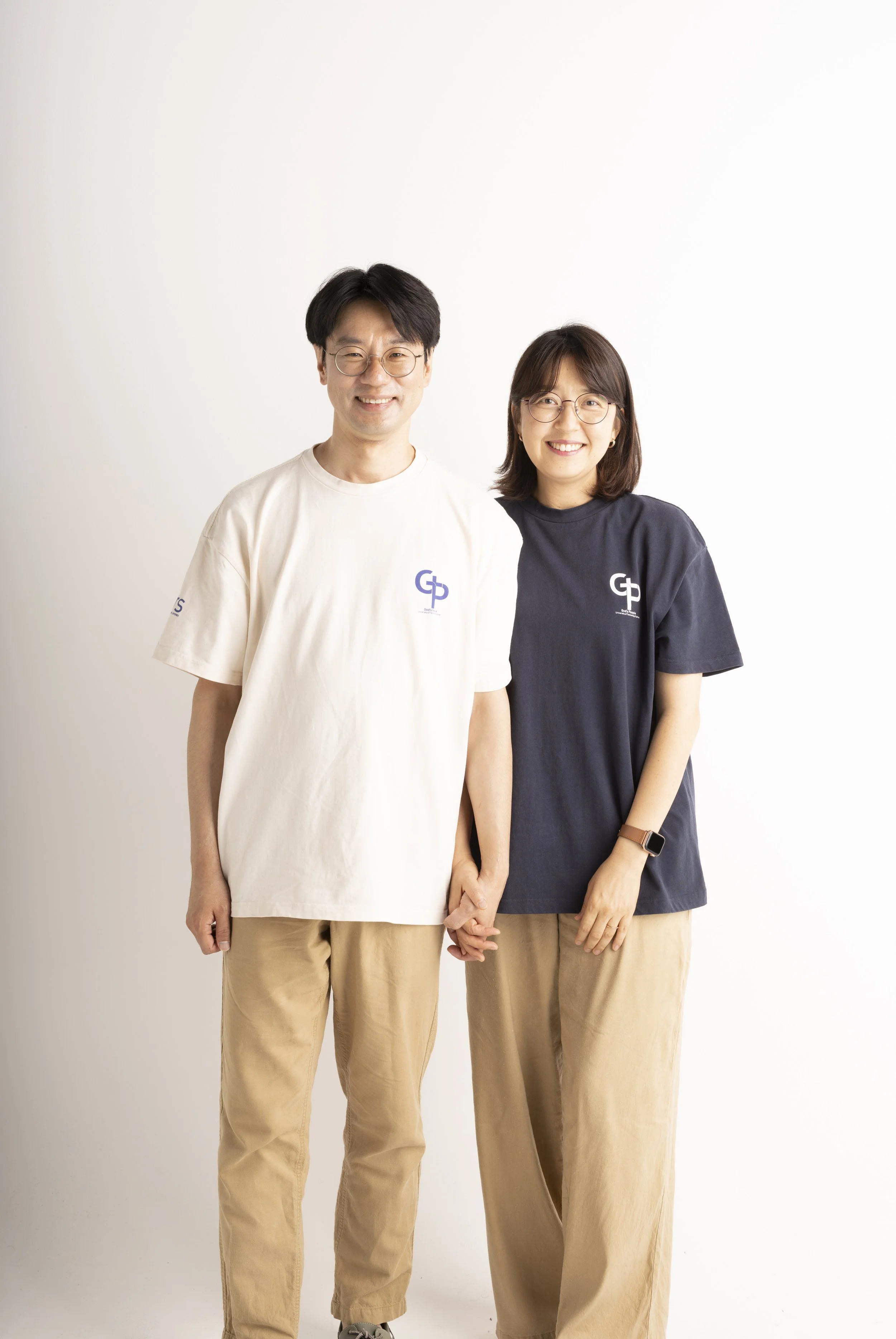 A smiling man and woman standing together, holding hands, wearing matching t-shirts with a logo, and beige pants against a plain white background.