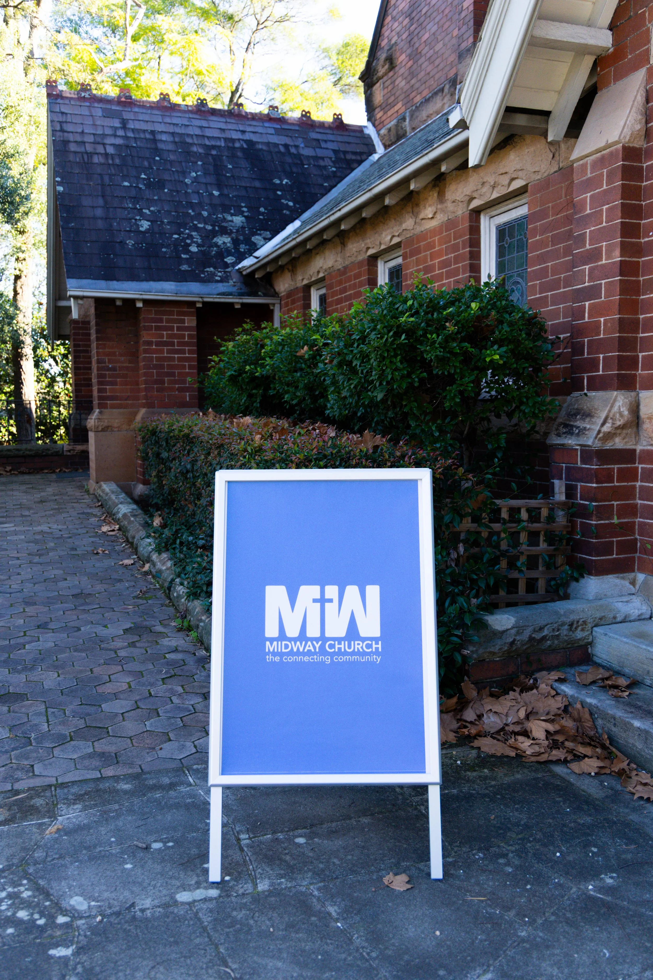Sign for Midway Church with the slogan 'the connecting community' in front of a brick building with windows and greenery.