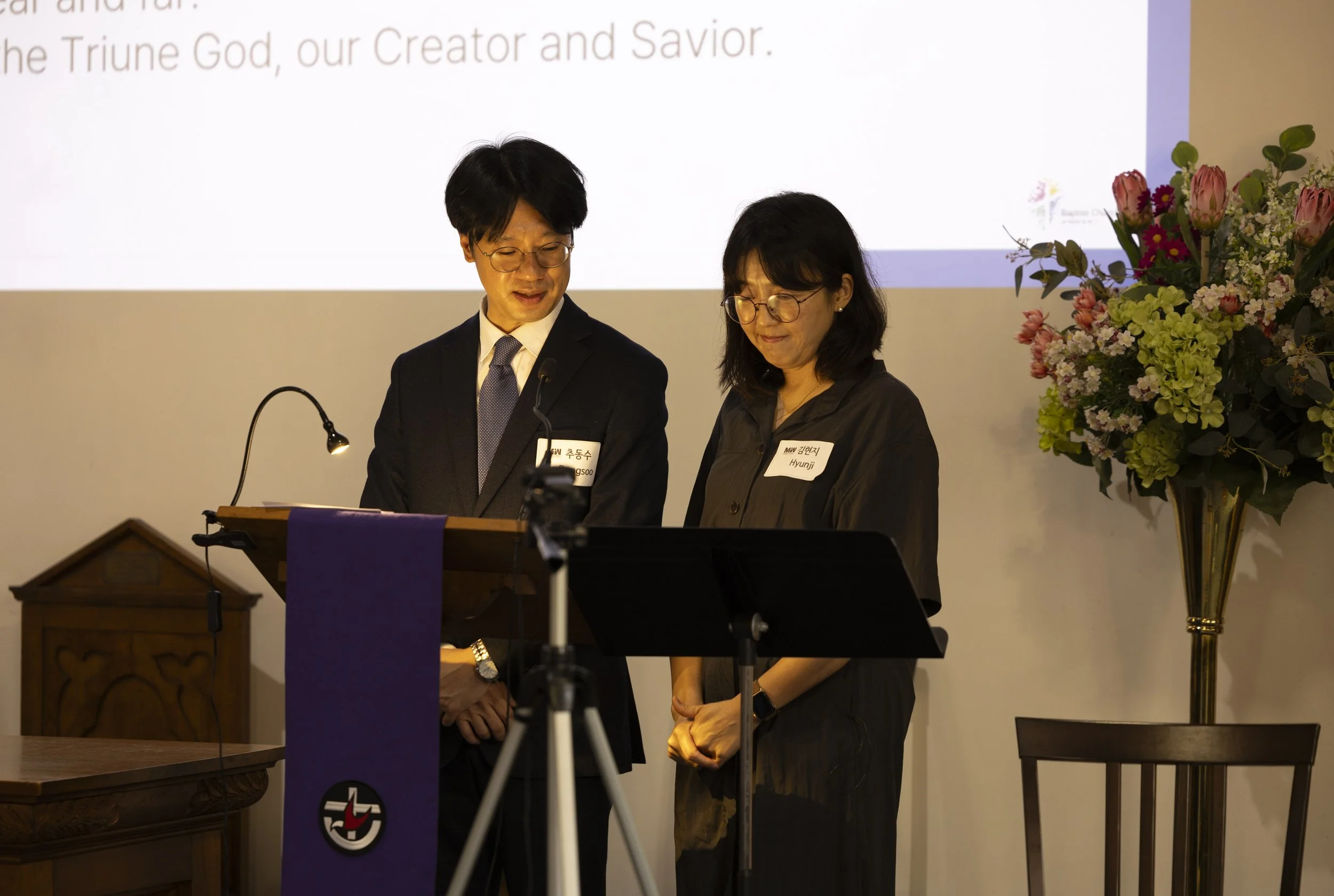 Two people standing at a podium in front of a church setting, a floral arrangement, and a large screen displaying partially visible text.