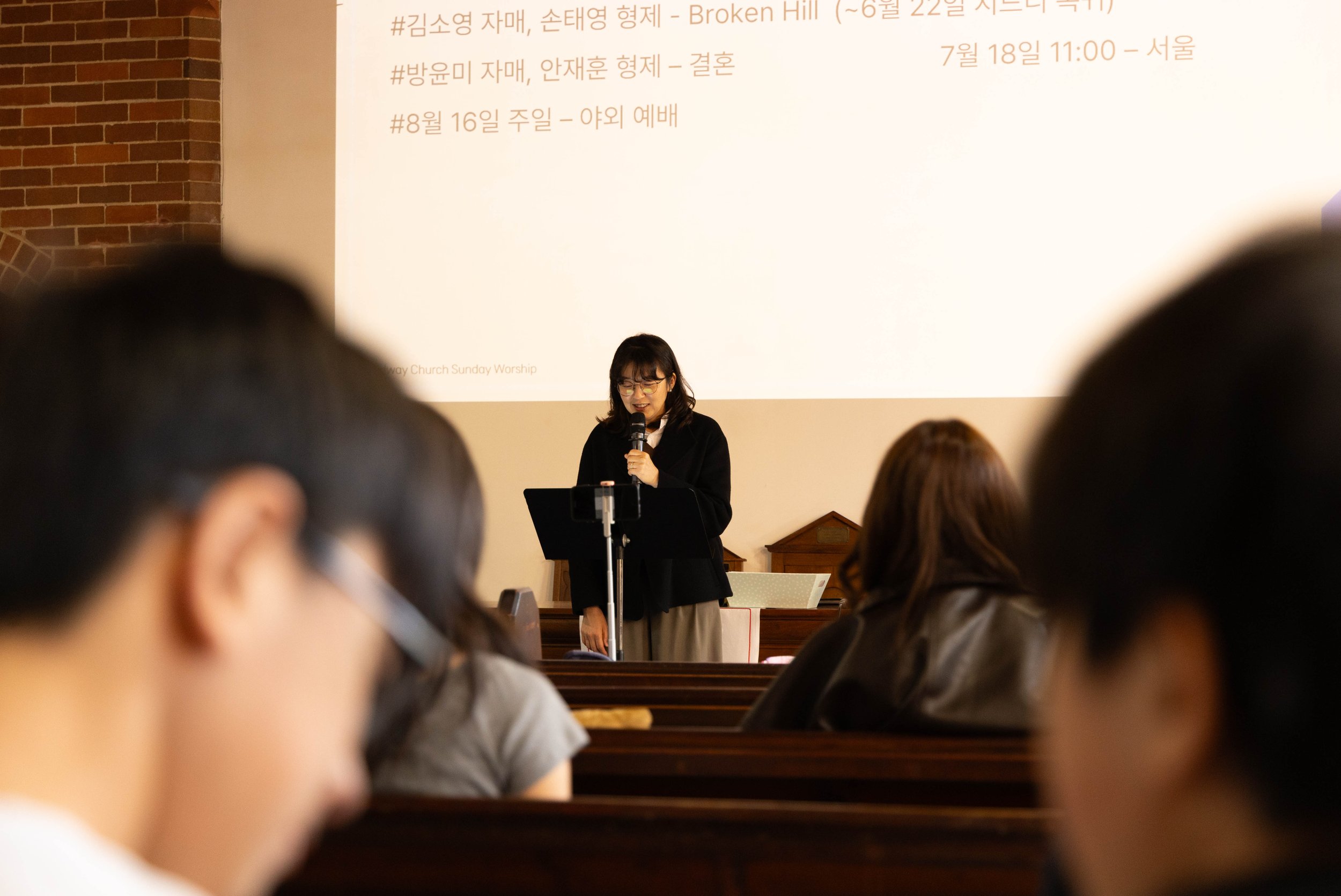 A woman with glasses speaking into a microphone at a podium in front of an audience in a church or event hall, with a large screen displaying text behind her. Audience members are seen from the back, with heads and shoulders visible.