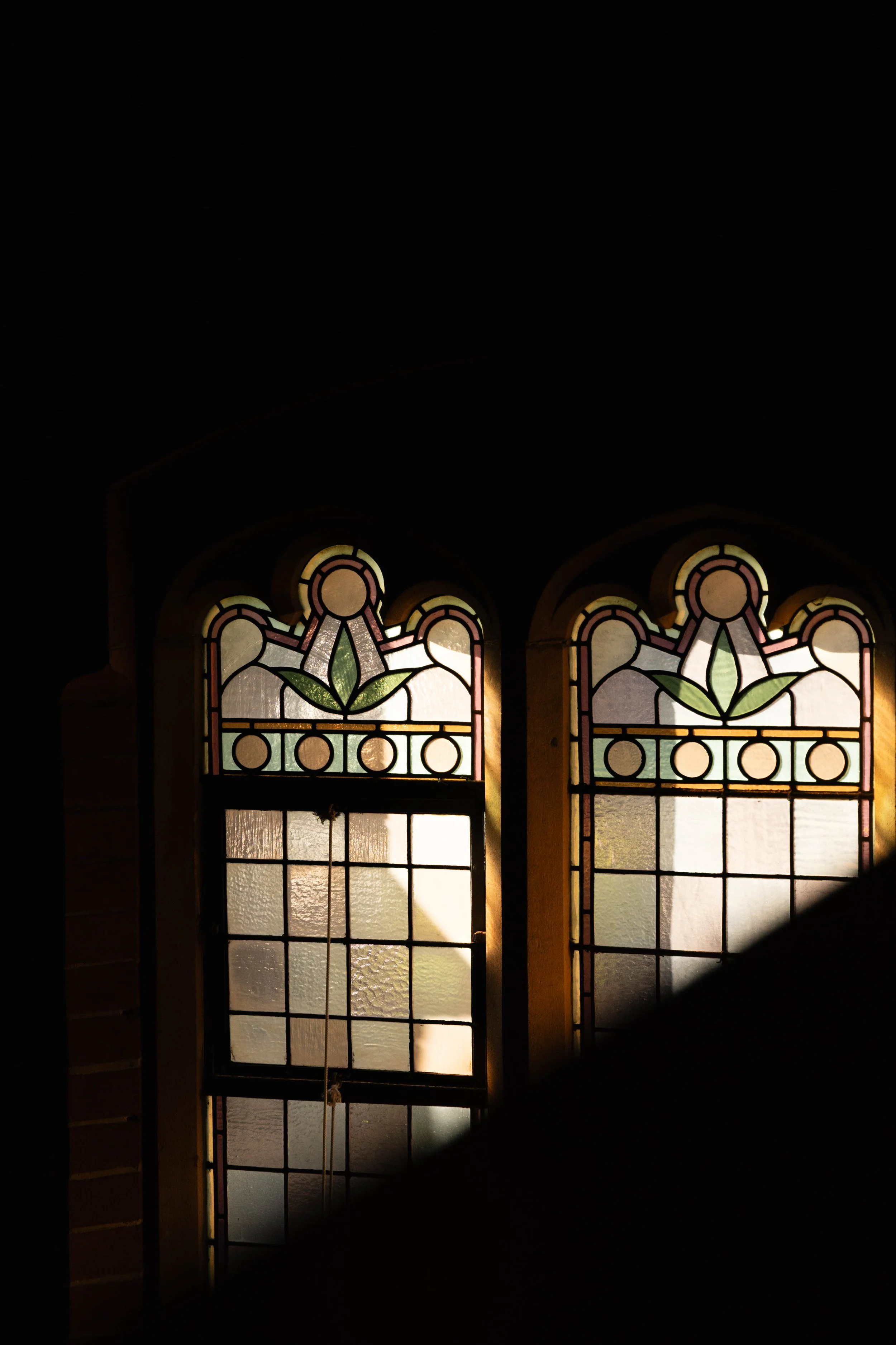 Two decorative stained glass windows with green leaf patterns and geometric designs, illuminated by sunlight, set in a dark room.