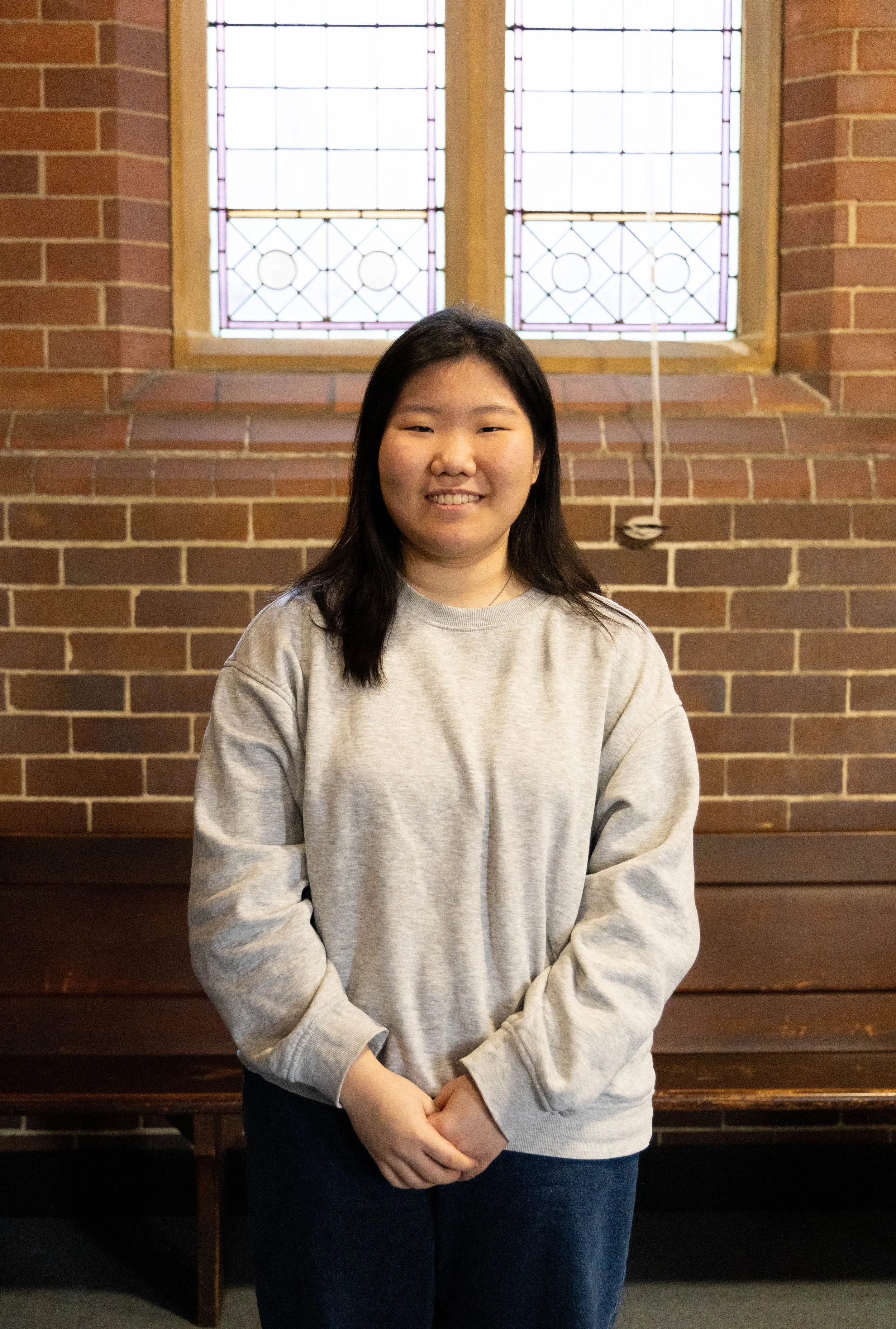 A young woman with long dark hair smiling, wearing a gray sweatshirt, standing in front of a brick wall with stained glass windows.
