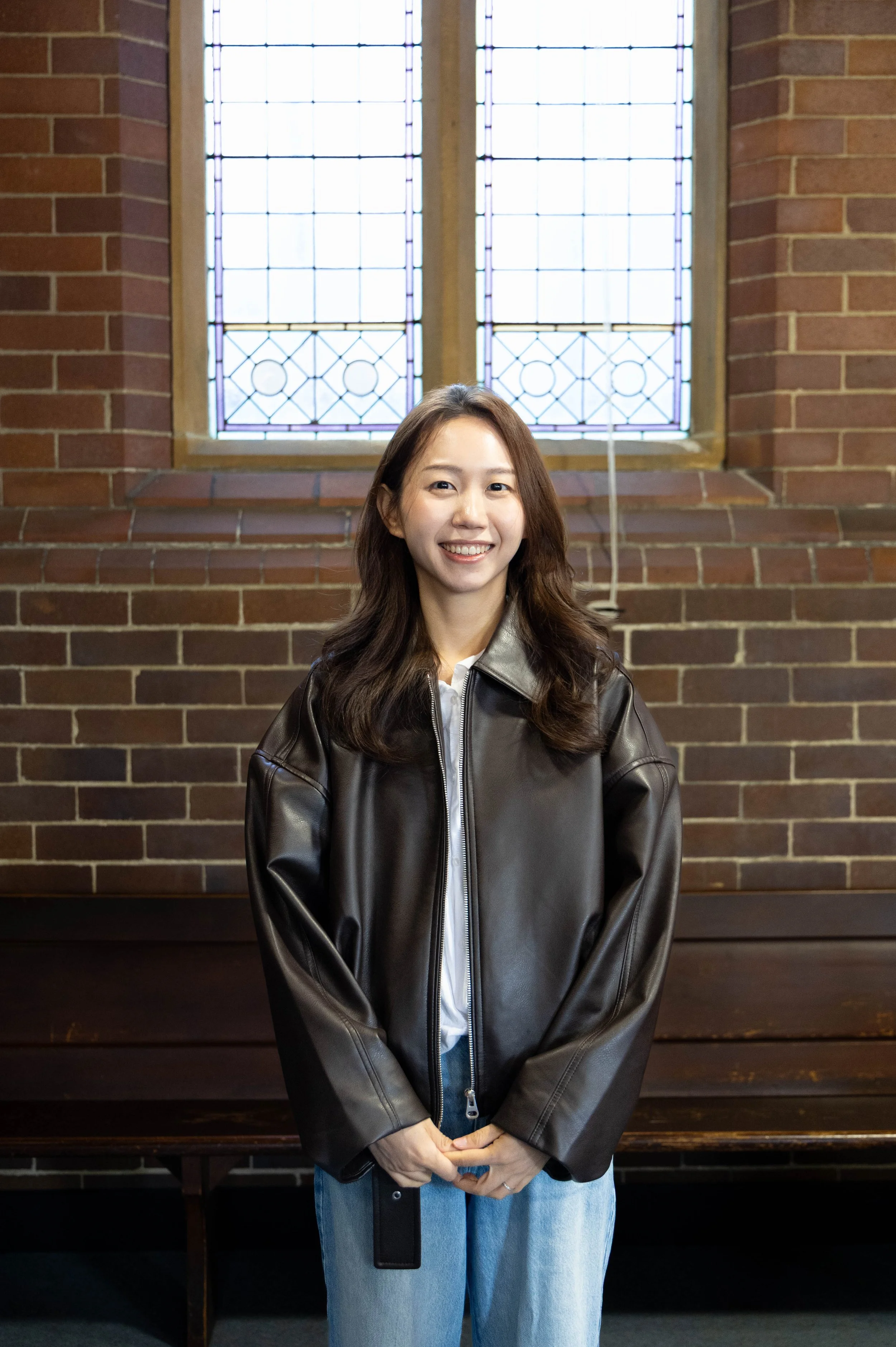 A young woman with long wavy hair, smiling, standing indoors in front of a brick wall and stained glass window, wearing a black leather jacket and blue jeans.