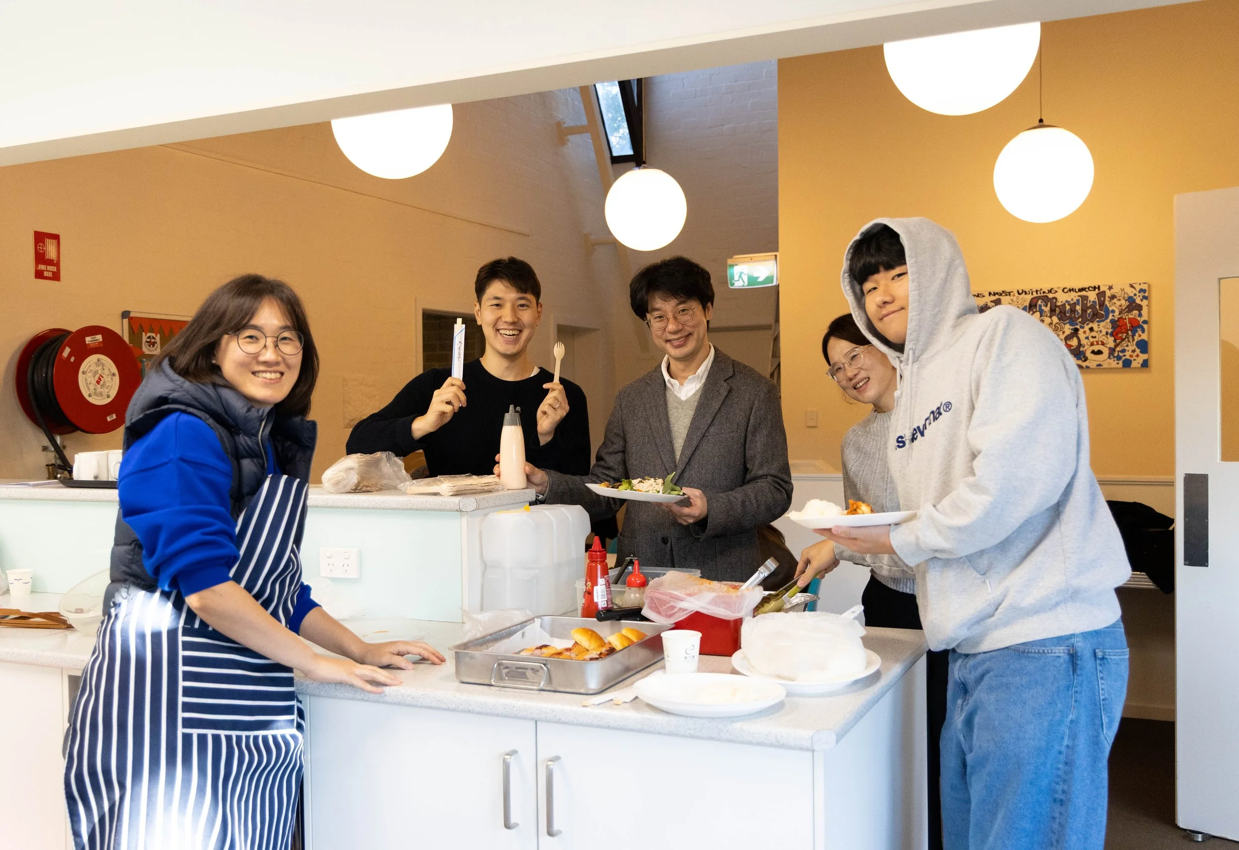 A group of five people gathered around a kitchen counter with food, smiling at the camera. The group includes two women and three men, one of whom is wearing a hoodie. The kitchen has hanging round lights, a fire alarm, and colorful wall decorations.