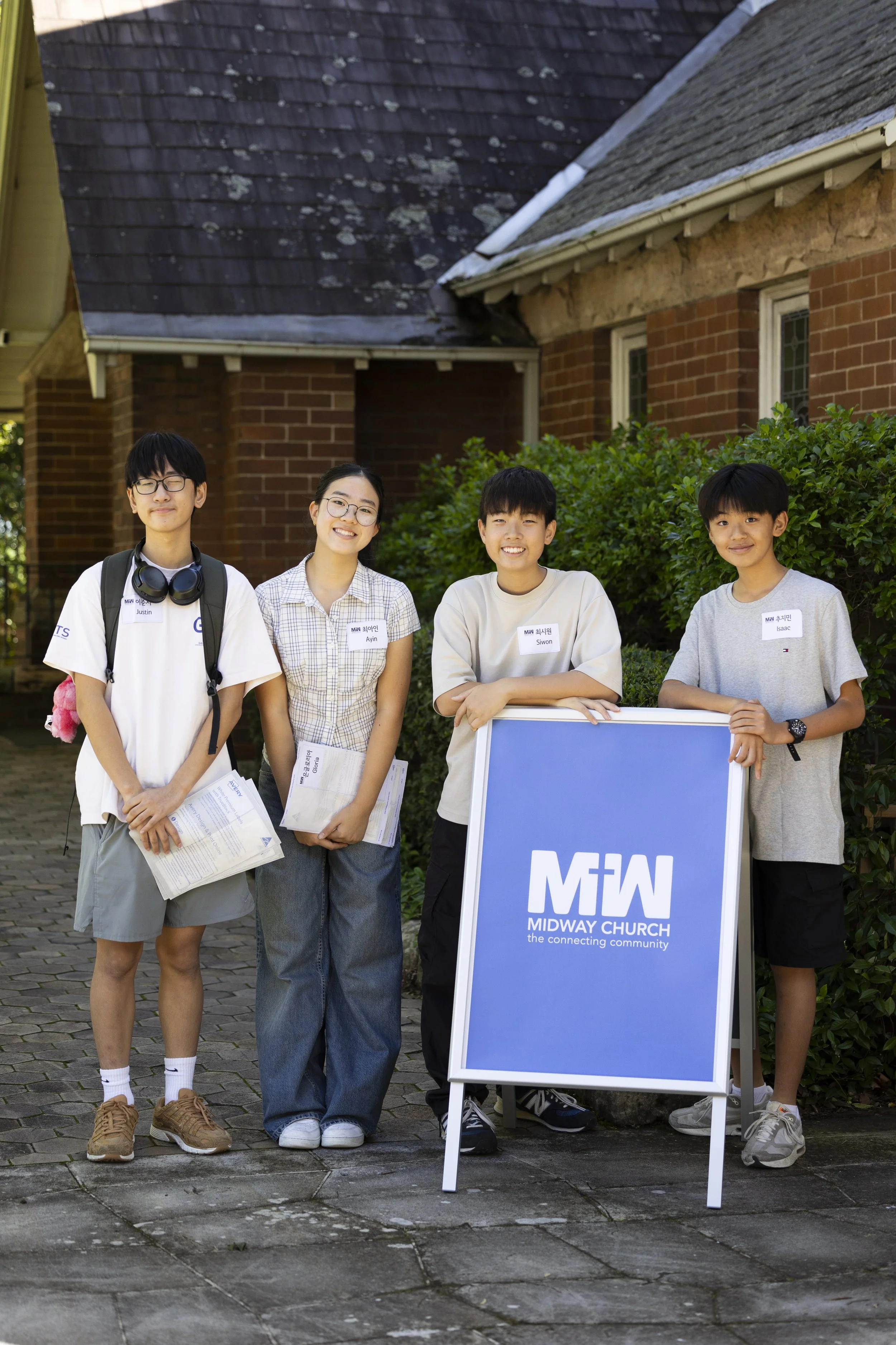 Four young people standing outdoors in front of a brick building and green shrubbery, smiling at the camera. One is holding a sign with the logo of Midway Church.