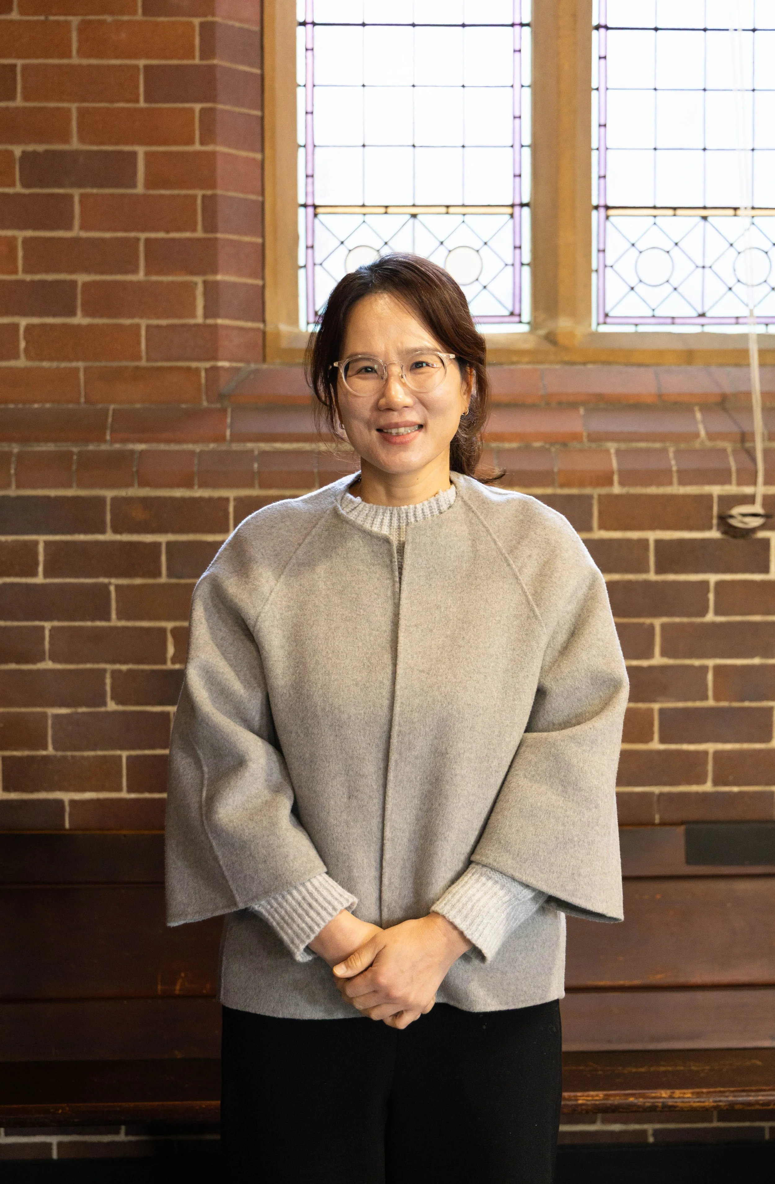 A woman with glasses wearing a gray coat, standing in front of a brick wall and stained glass windows.
