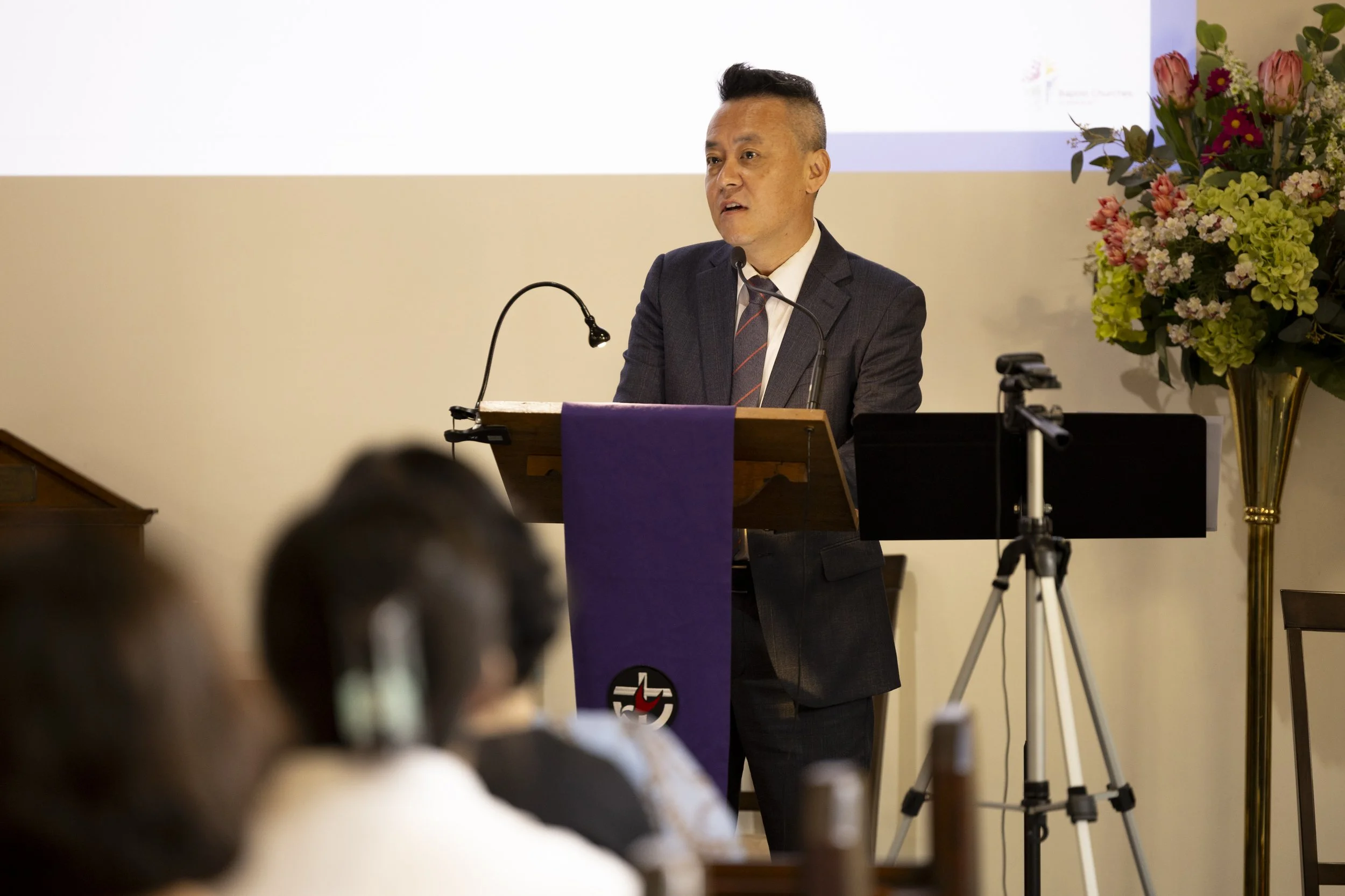Man in dark suit and striped tie speaking at a podium during a presentation, with flowers on a stand to his right and an audience in the foreground.