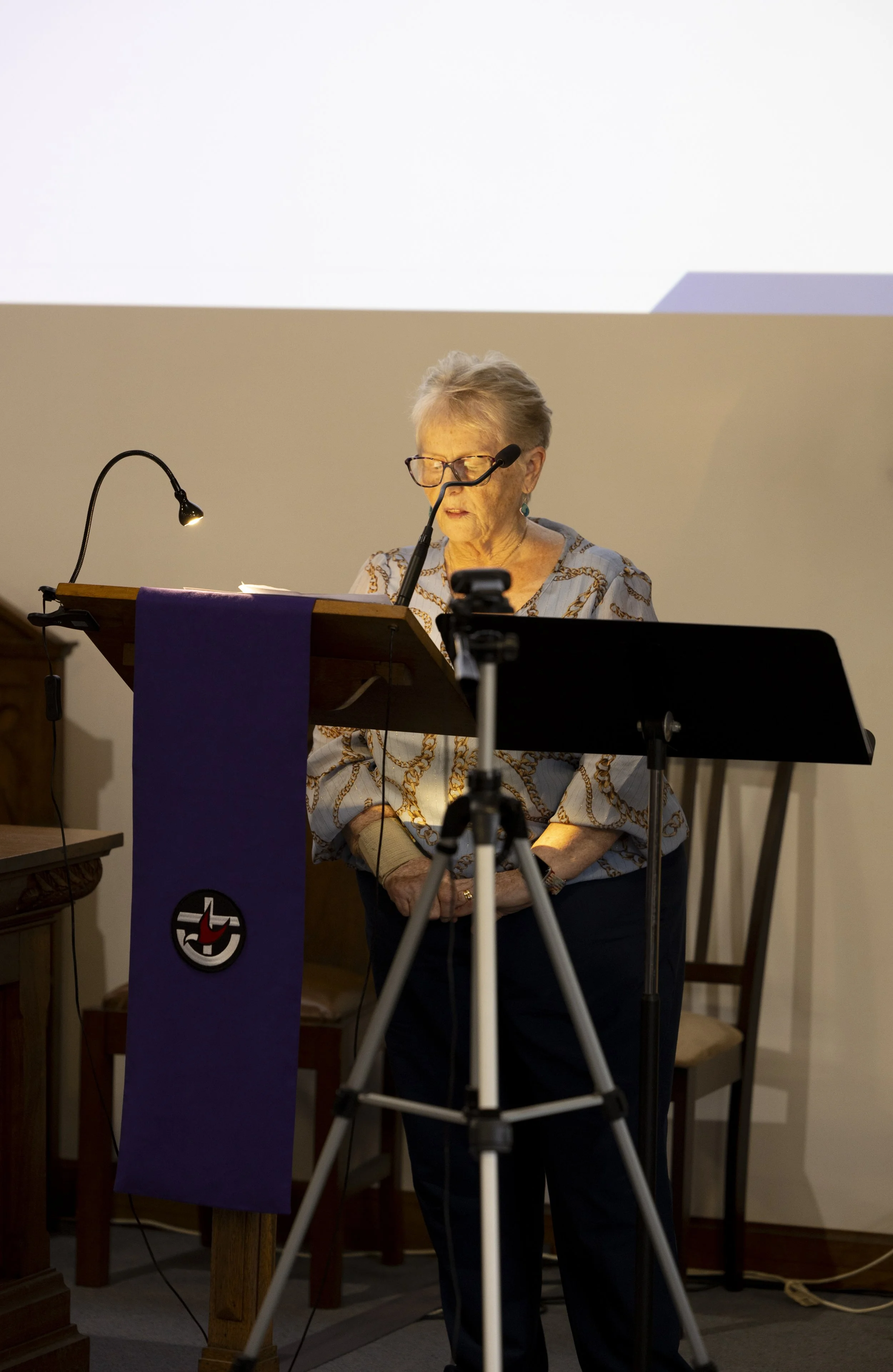 An elderly woman with glasses and gray hair standing at a lectern, reading or speaking, in a room with a large white projector screen behind her. The lectern has a purple cloth with a logo, and there is a microphone and reading lamp on it. The woman 