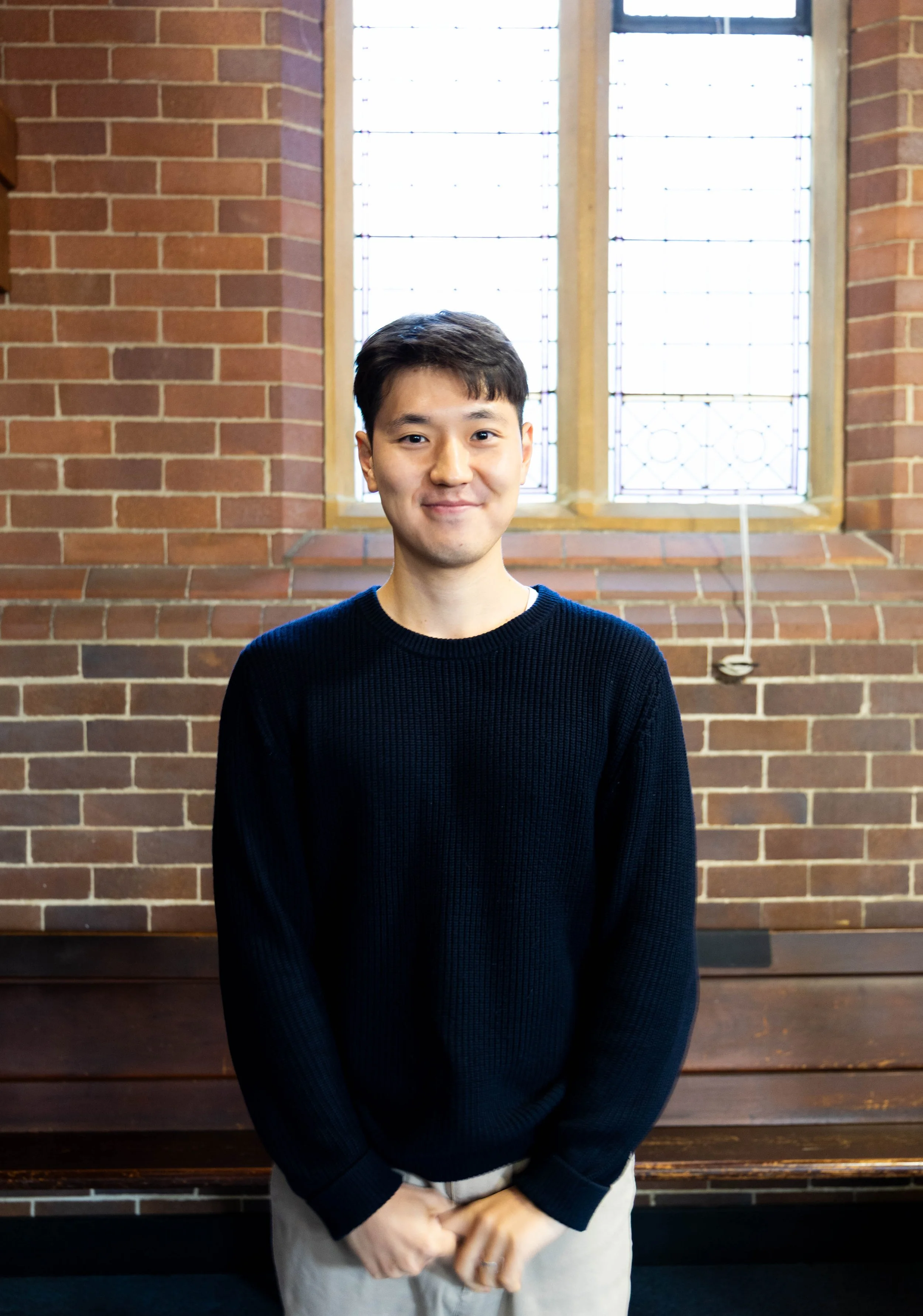 A young man with dark hair and a slight smile standing in front of a brick wall with large windows behind him. He is wearing a black sweater and light-colored pants, with hands folded in front.