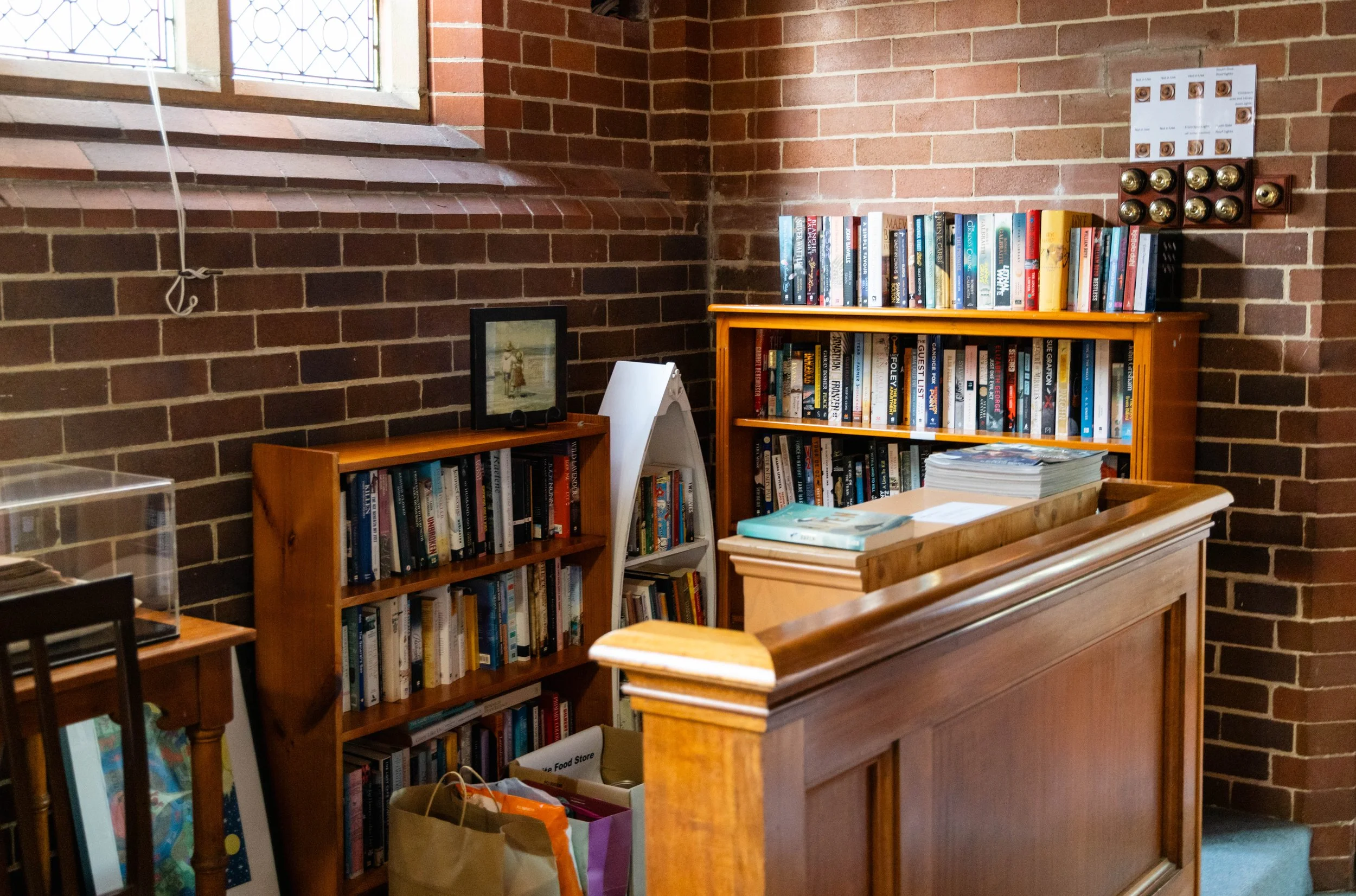 Bookshelves filled with books against brick walls in a cozy corner of a room, with a window overhead and some packaged items in the foreground.