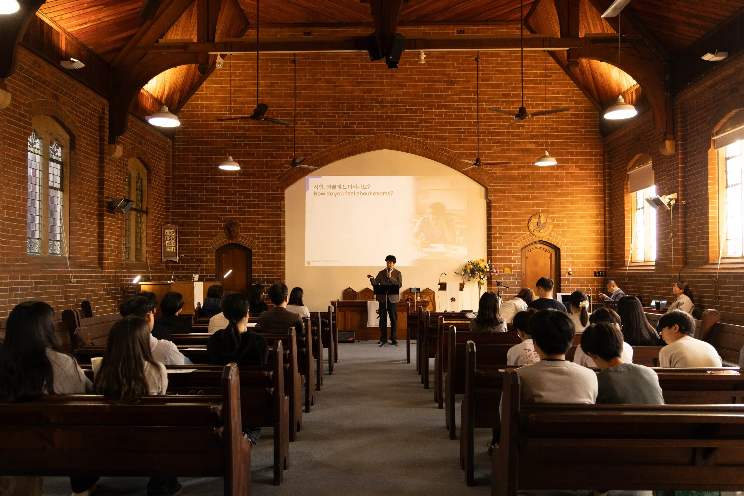 A speaker giving a presentation at the front of a church or lecture hall filled with seated audience. The room has brick walls, stained glass windows, and a high wooden arched ceiling.