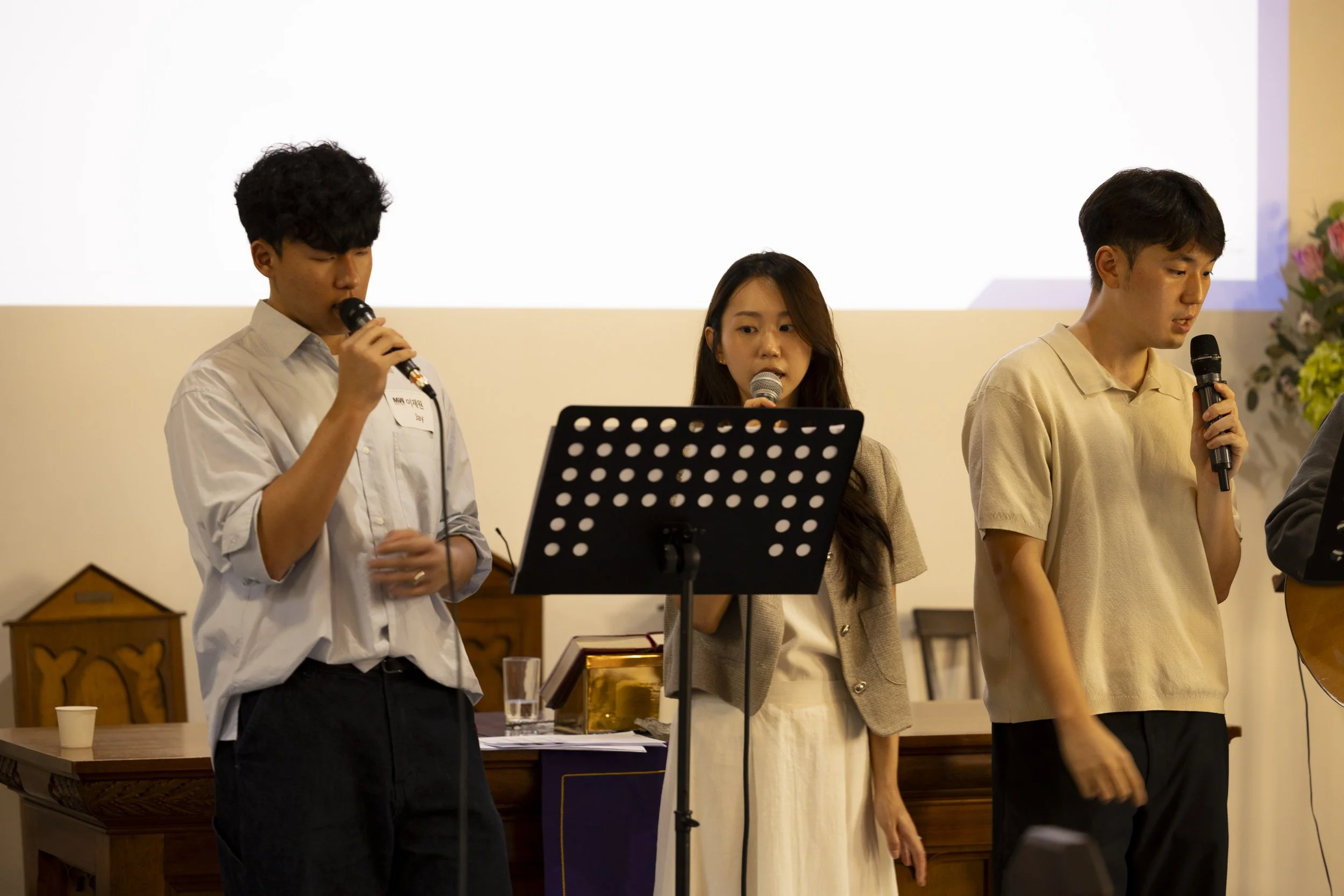 Three young people standing in a row, each holding a microphone, in what appears to be a formal or semi-formal event. The young man on the left wears a white shirt, the young woman in the middle wears a beige jacket, and the young man on the right we