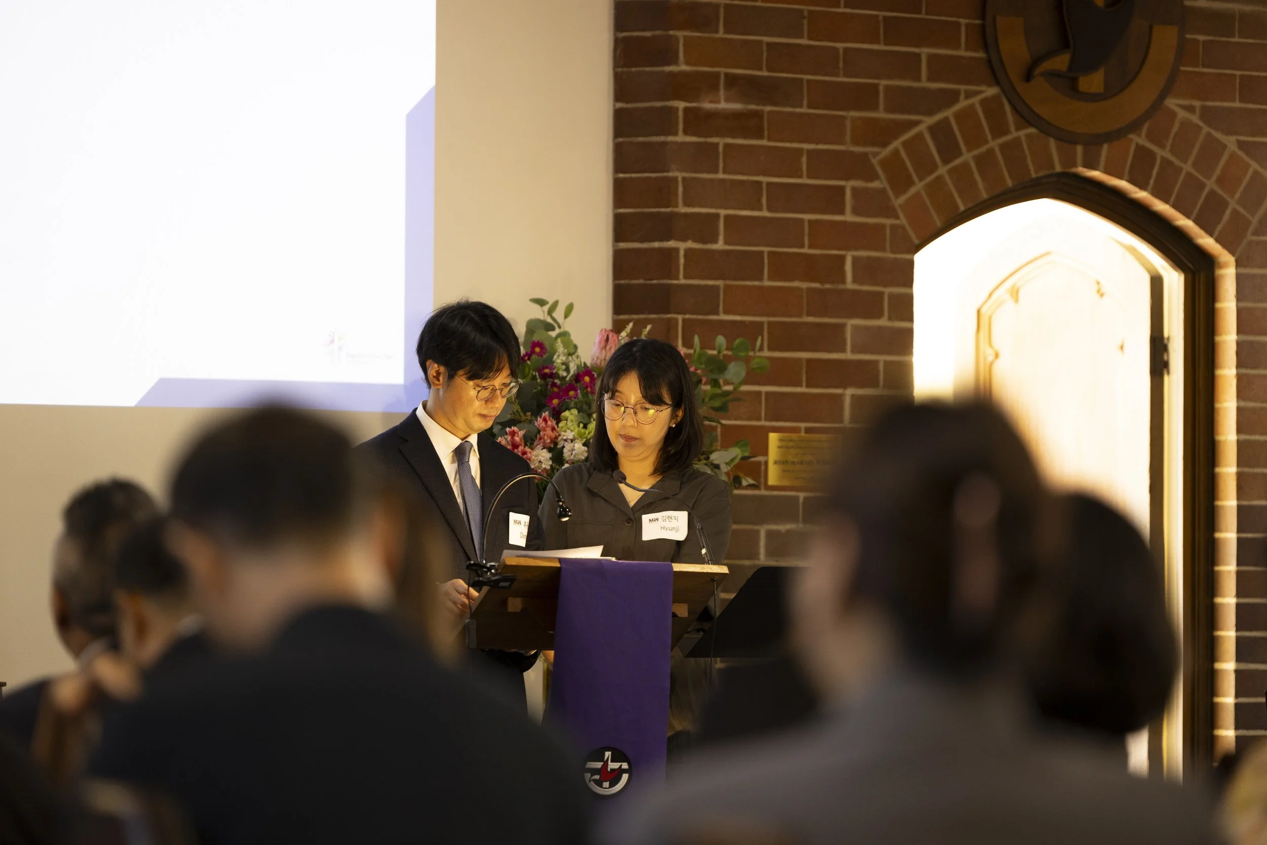 Two individuals stand at a podium during a formal event, with a brick wall in the background and a floral arrangement to the side. The audience is visible in the foreground.