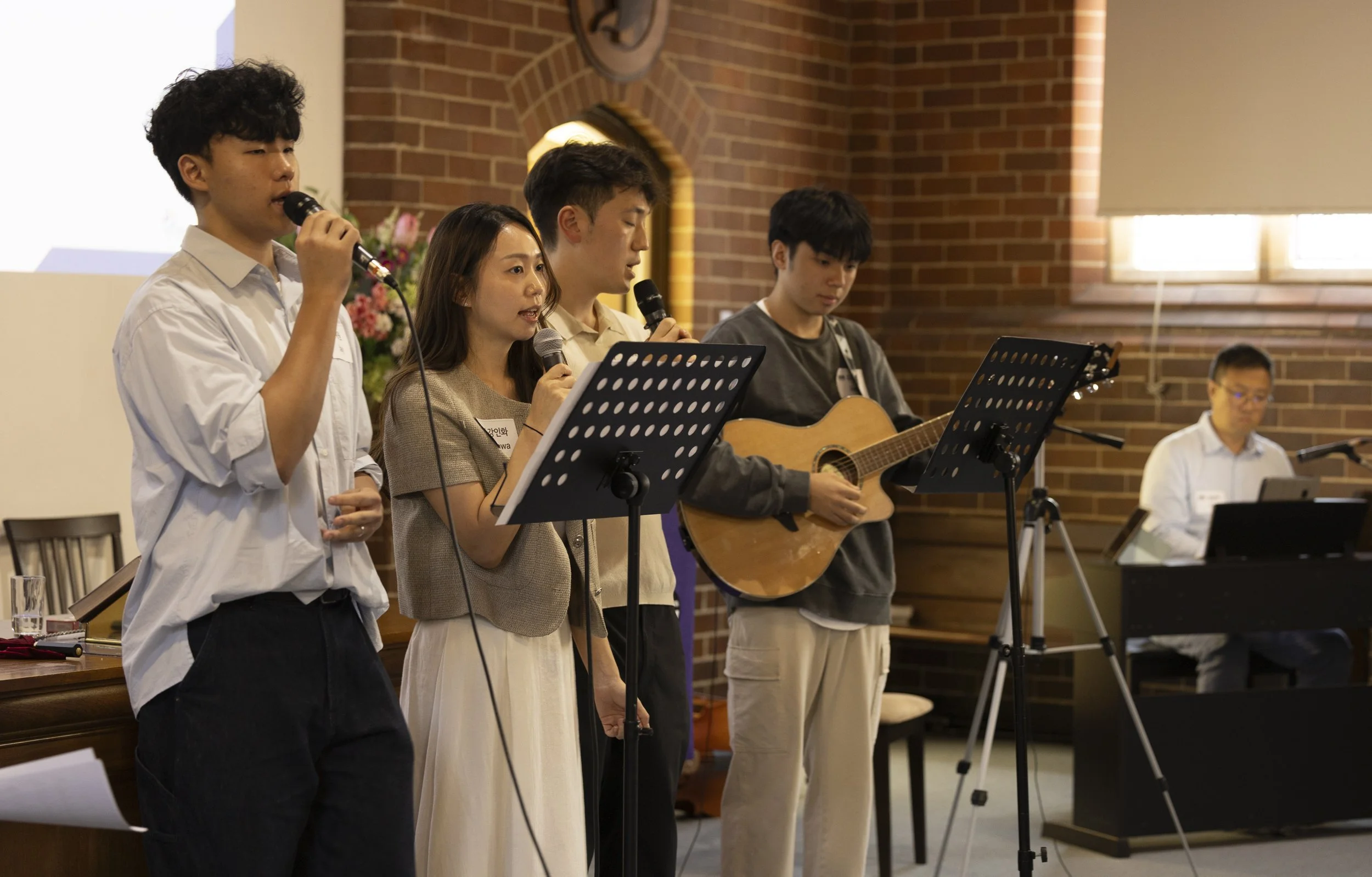 Group of five young people performing music together in a church or community hall. Two are singing into microphones, one is playing an acoustic guitar, and another is playing keyboard. They are standing in front of a brick wall with a window and a floral arrangement.