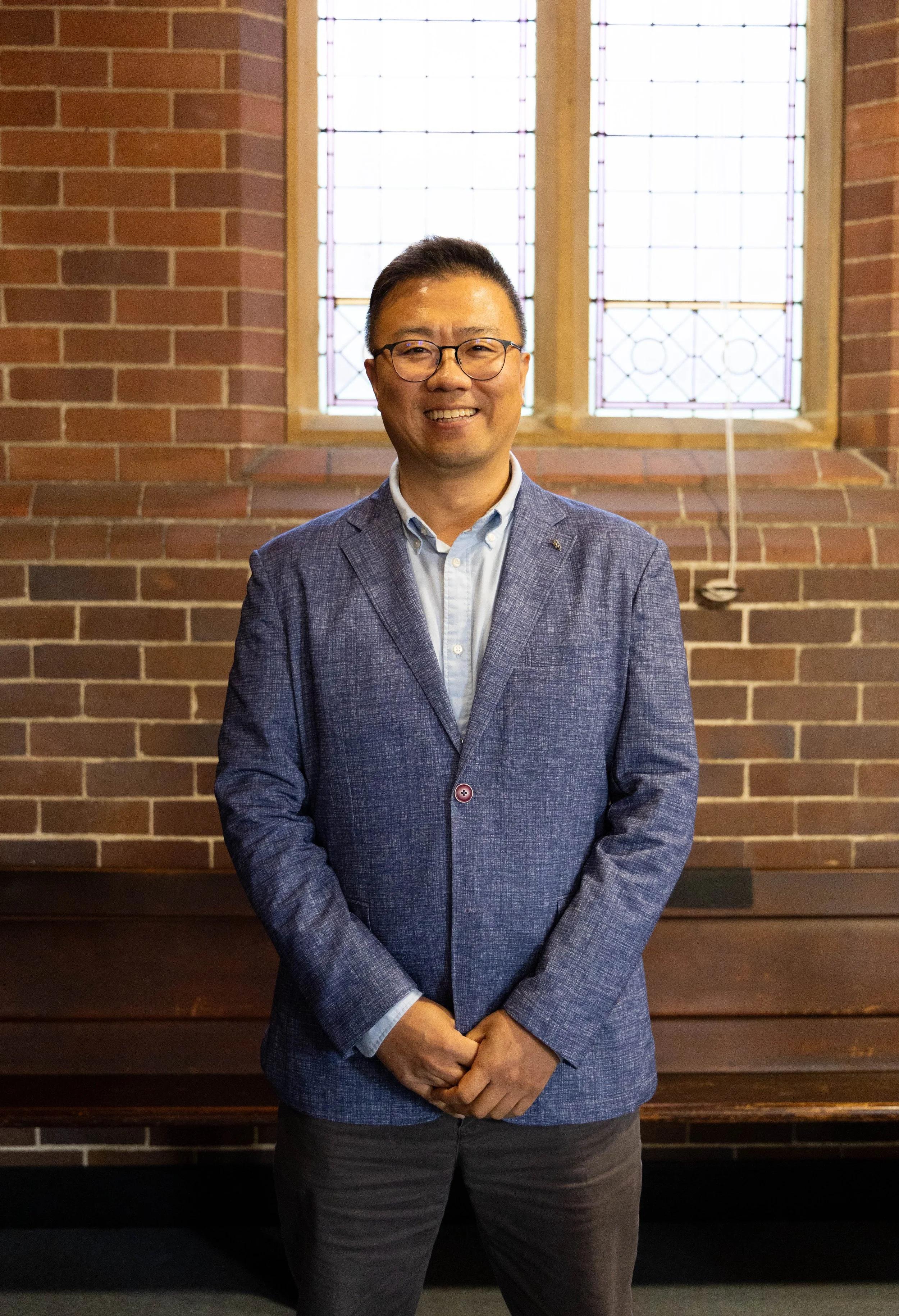 A smiling man with glasses wearing a blue blazer and light blue shirt, standing in front of a brick wall with stained glass windows.