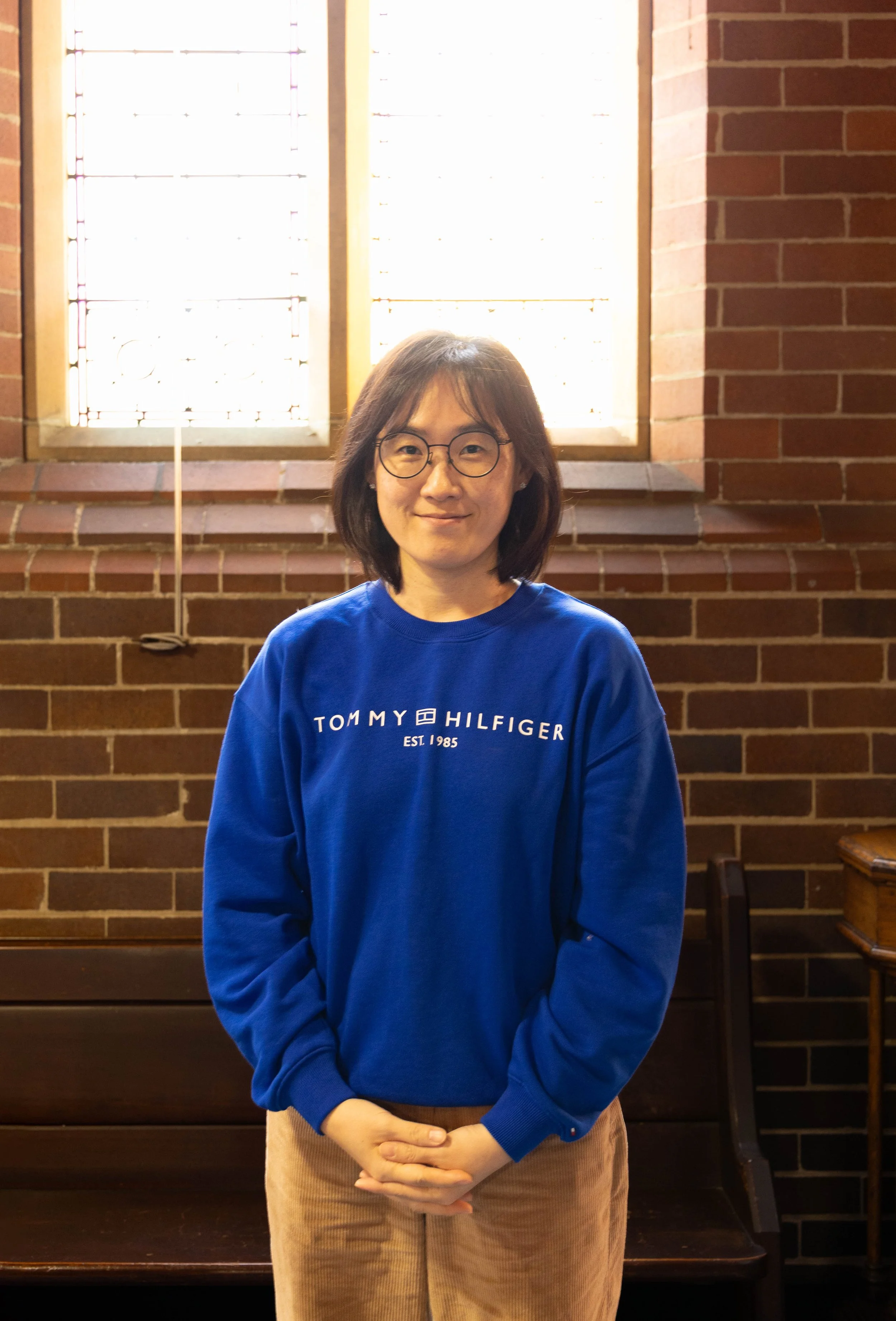 A woman with glasses, wearing a blue Tommy Hilfiger sweatshirt and beige pants, standing indoors against a brick wall with a window behind her.