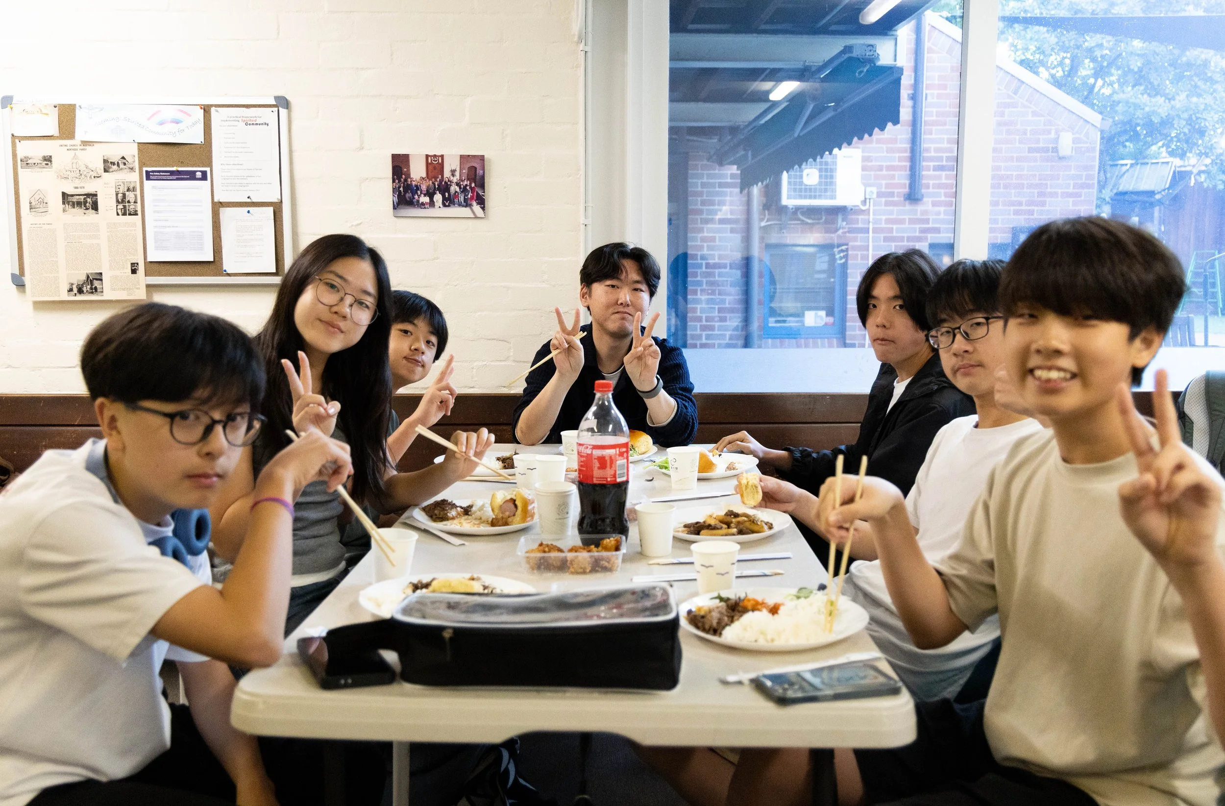 Group of young people sitting at a table enjoying a meal, some making peace signs and smiling.