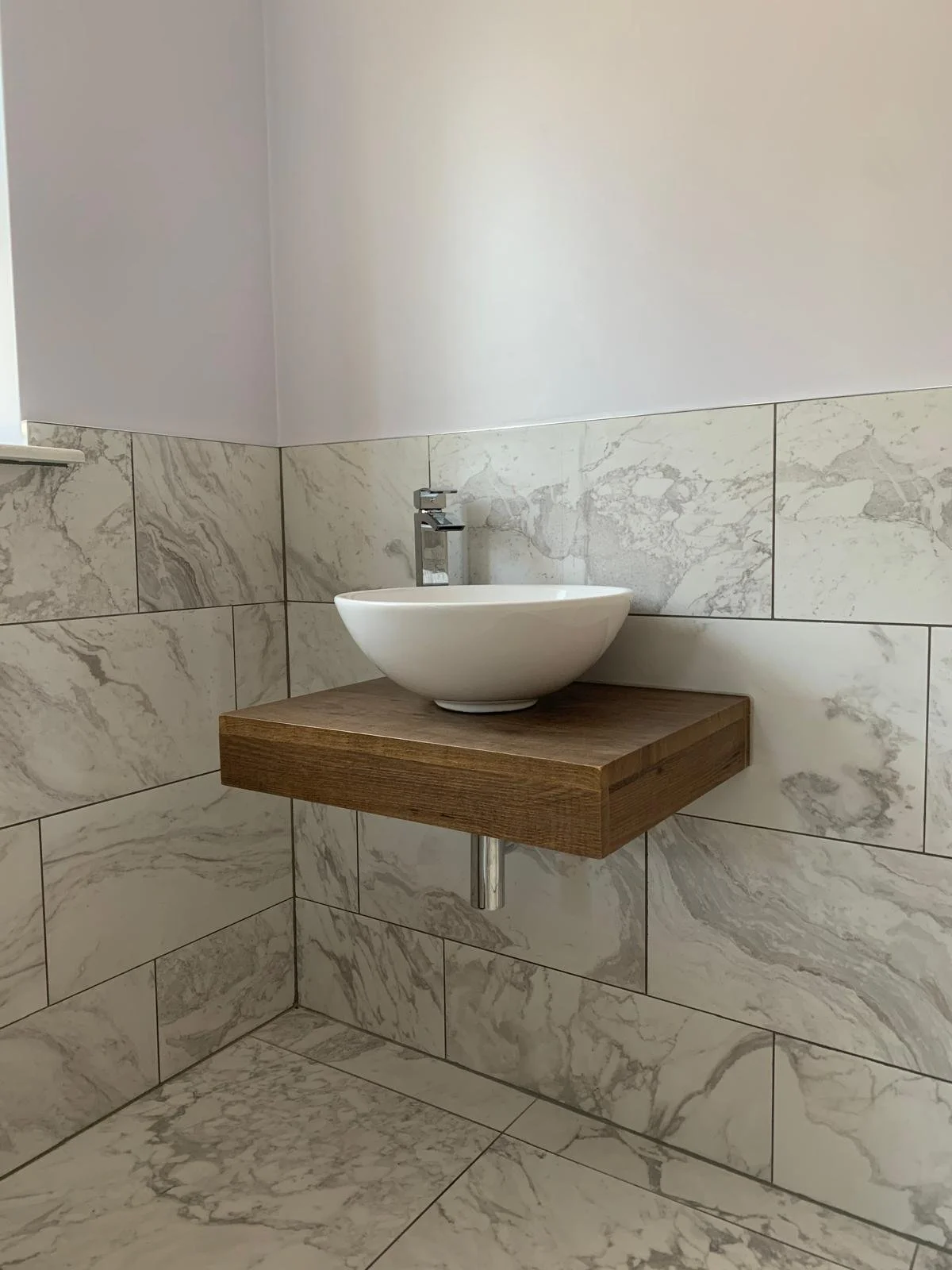 Bathroom corner with marble tiled walls and floor, a floating wooden sink with a white bowl-shaped vessel sink, and a modern chrome faucet.