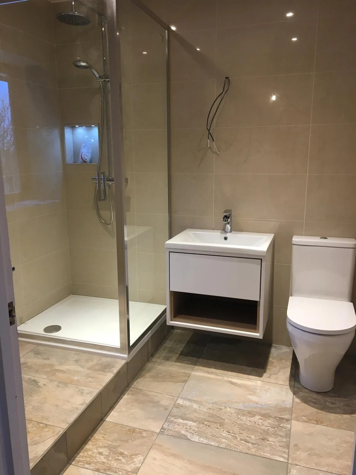 Bathroom with shower stall, white sink vanity, and toilet, beige tiled walls and floor, some electrical wires hanging from the wall.