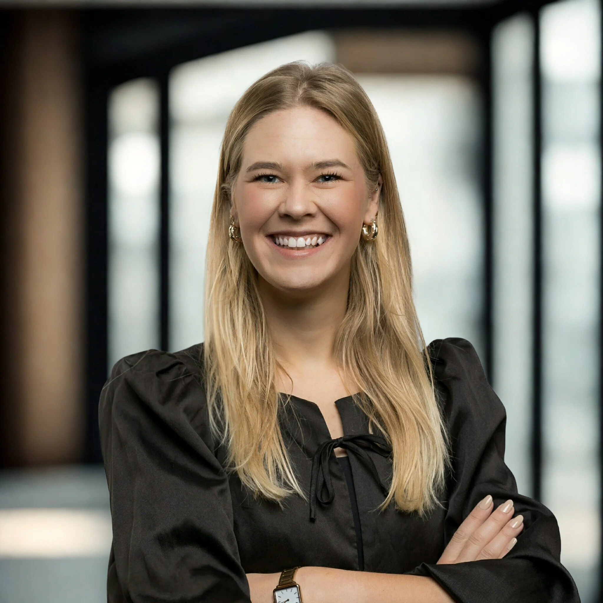 Portret van een lachende jonge vrouw met blond haar in een zwarte blouse, gestoken armen, in een moderne kantooromgeving.