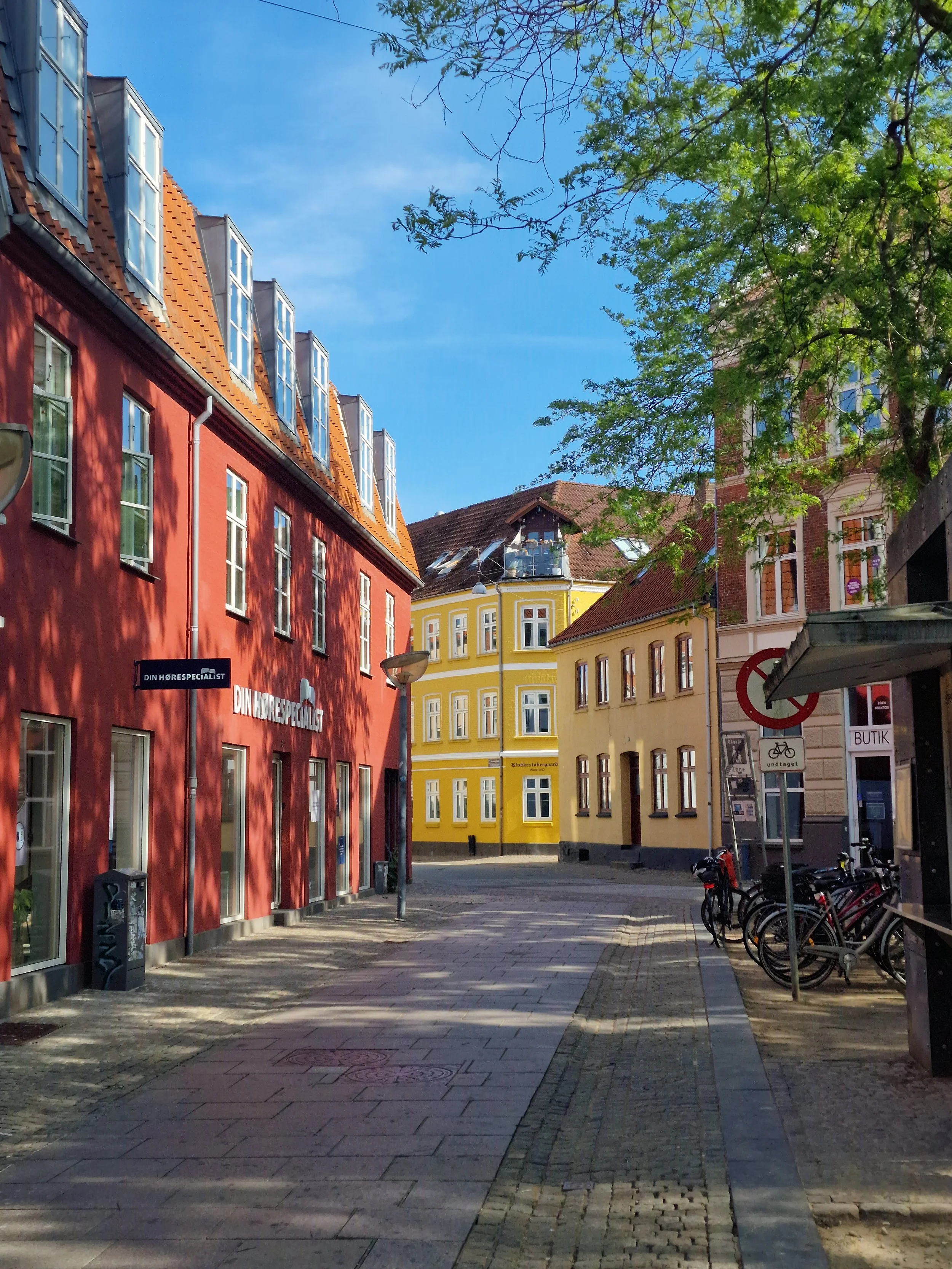 Een straat met kleurrijke gebouwen, voornamelijk rood, geel en beige, onder een blauwe hemel met een paar wolken. Bomen met groene bladeren werpen schaduw op de straat. Fietsen staan geparkeerd aan de rechterkant.