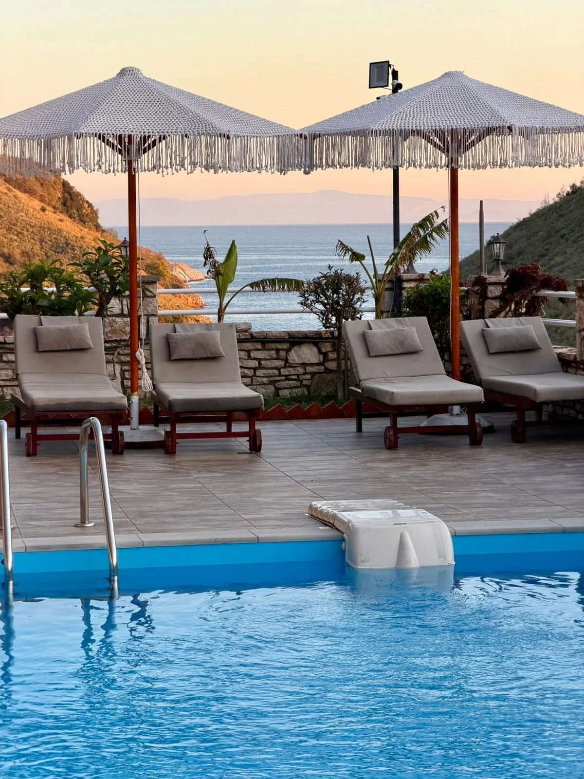 Uitzicht op een zwembad met ligstoelen en parasols, uitzicht op zee en bergen in de achtergrond tijdens zonsondergang.