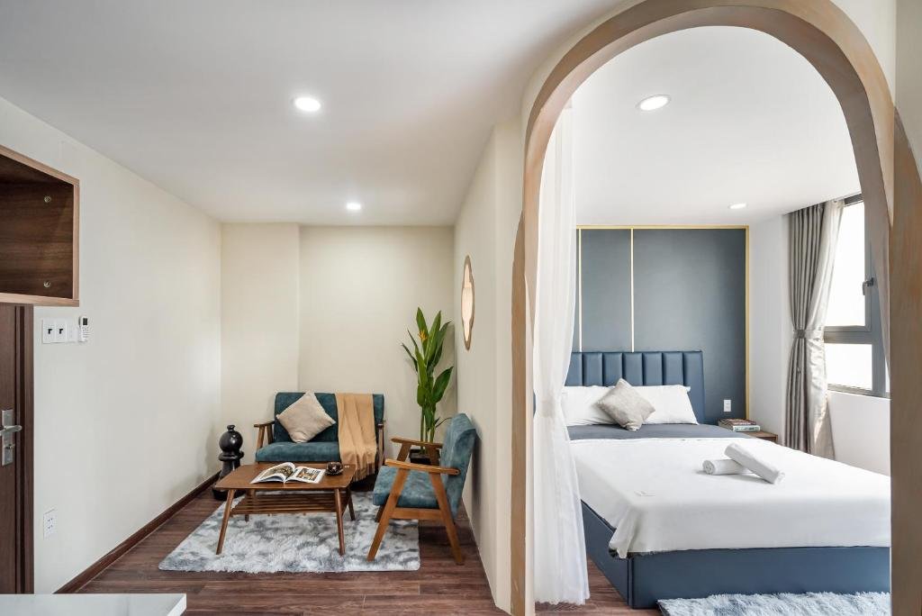 Een hotelkamer met een woonkamer en slaapkamer gescheiden door een open boog. De woonkamer heeft een vintagebankje, twee stoelen en een plant. De slaapkamer heeft een bed met wit beddengoed, een raam met gordijnen en een blauwe hoofdhoek.