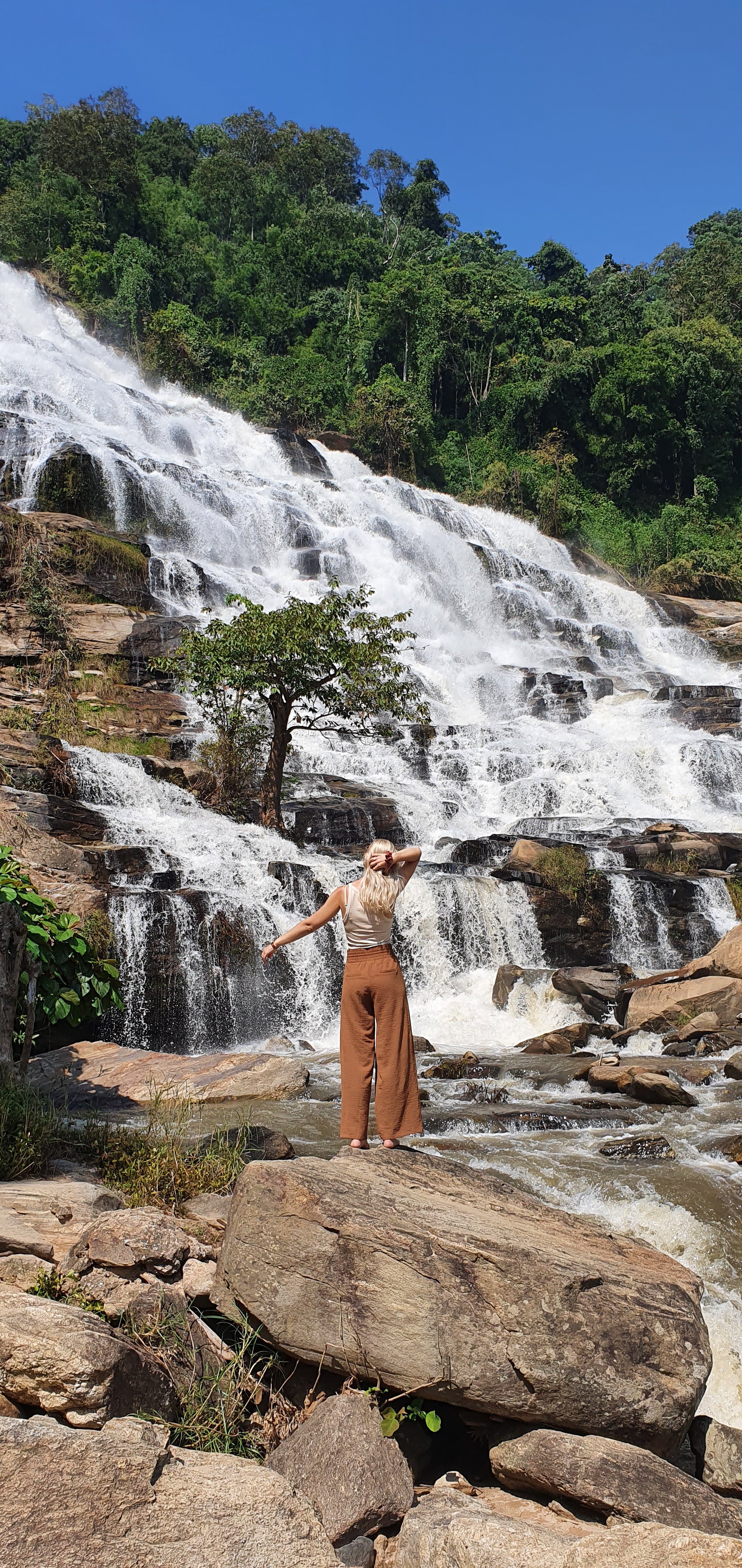 Een vrouw met lang blond haar, gedragen in een witte top en bruin broek, staat op rotsen naast een waterval, omgeven door groen bos en blauwe hemel.