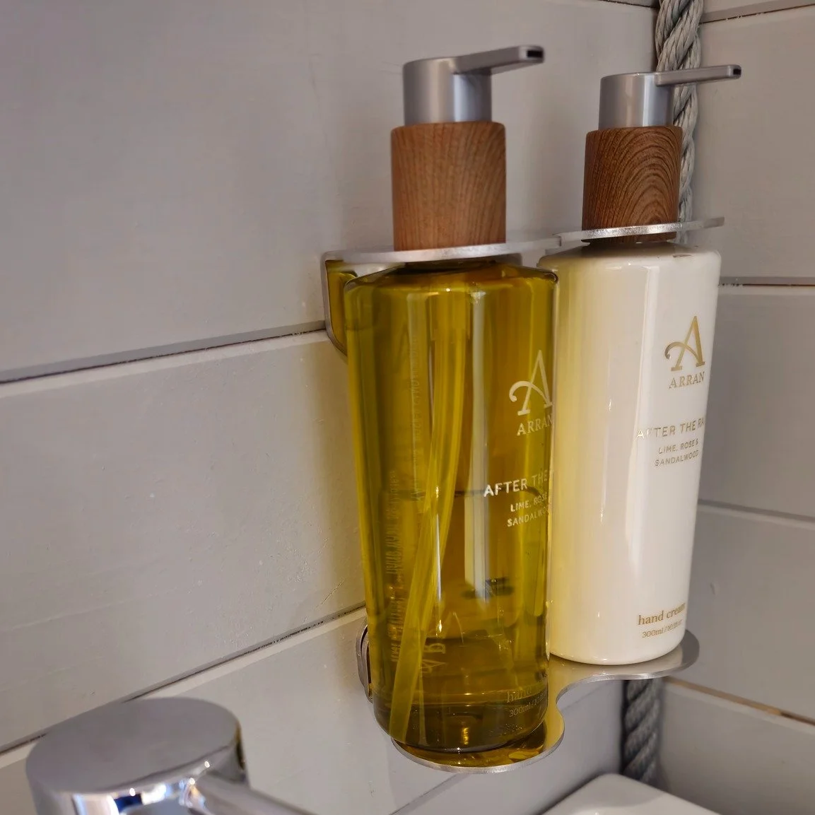 Two bottles of shower products with wooden caps mounted on a bathroom wall, one containing yellow liquid and the other containing white cream, labeled with brand ABRAN and scent "LIME, ROSES & SANDALWOOD."
