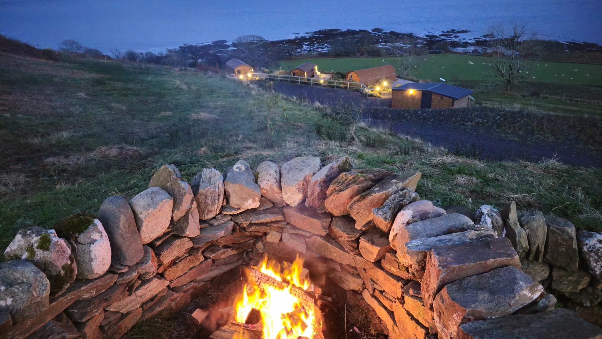 A stone fire pit with a blazing fire in front of a grassy hillside with houses and lights, landscape at dusk or early evening.